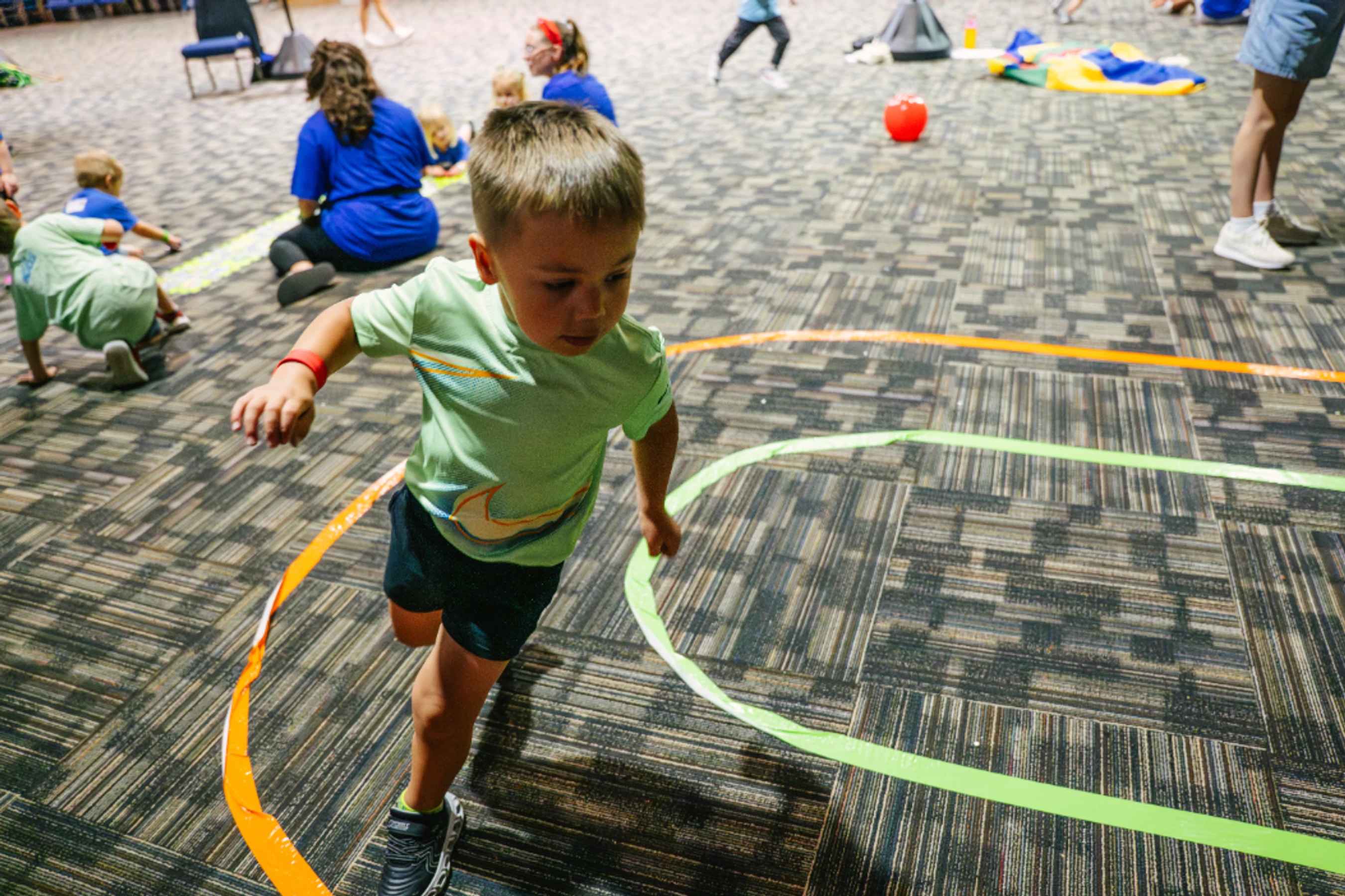 A young boy in a light green shirt runs along a track made of orange and green tape on a patterned carpet during an indoor group activity.