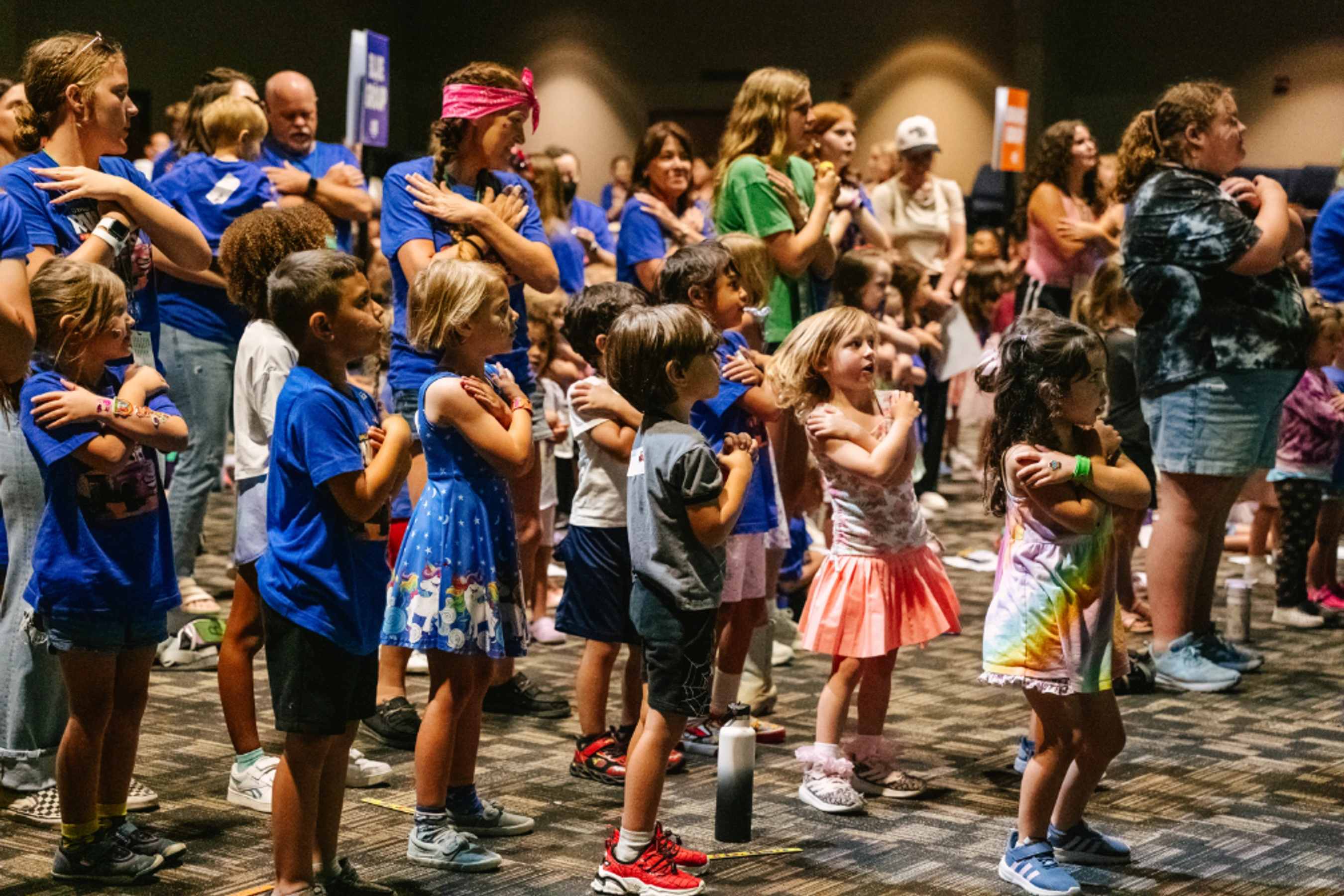 A large group of children and adults stand indoors with arms crossed over their chests, looking attentively to the side during a community gathering.