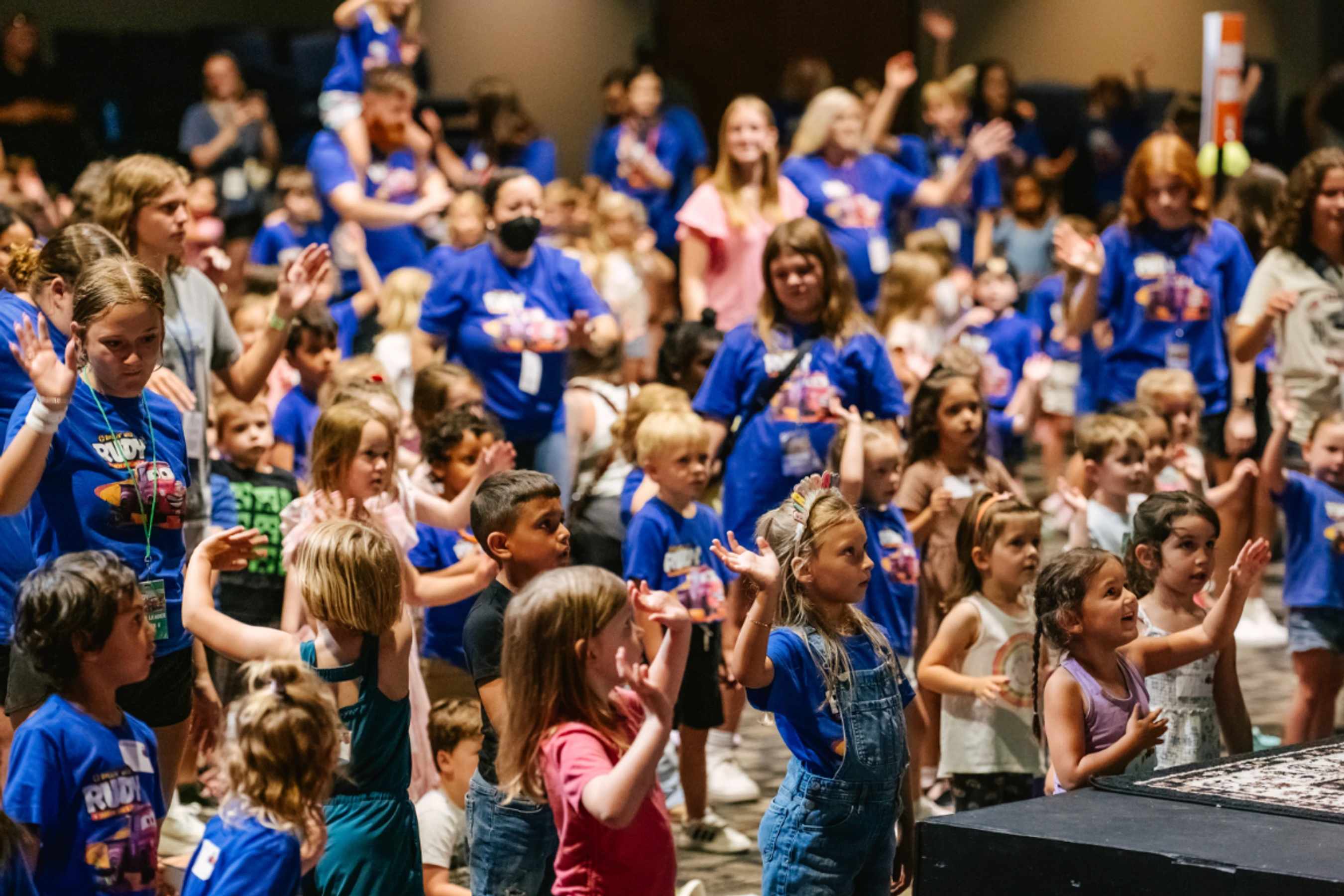 A large, diverse group of young children and adult leaders raise their hands together during an energetic assembly in an auditorium.