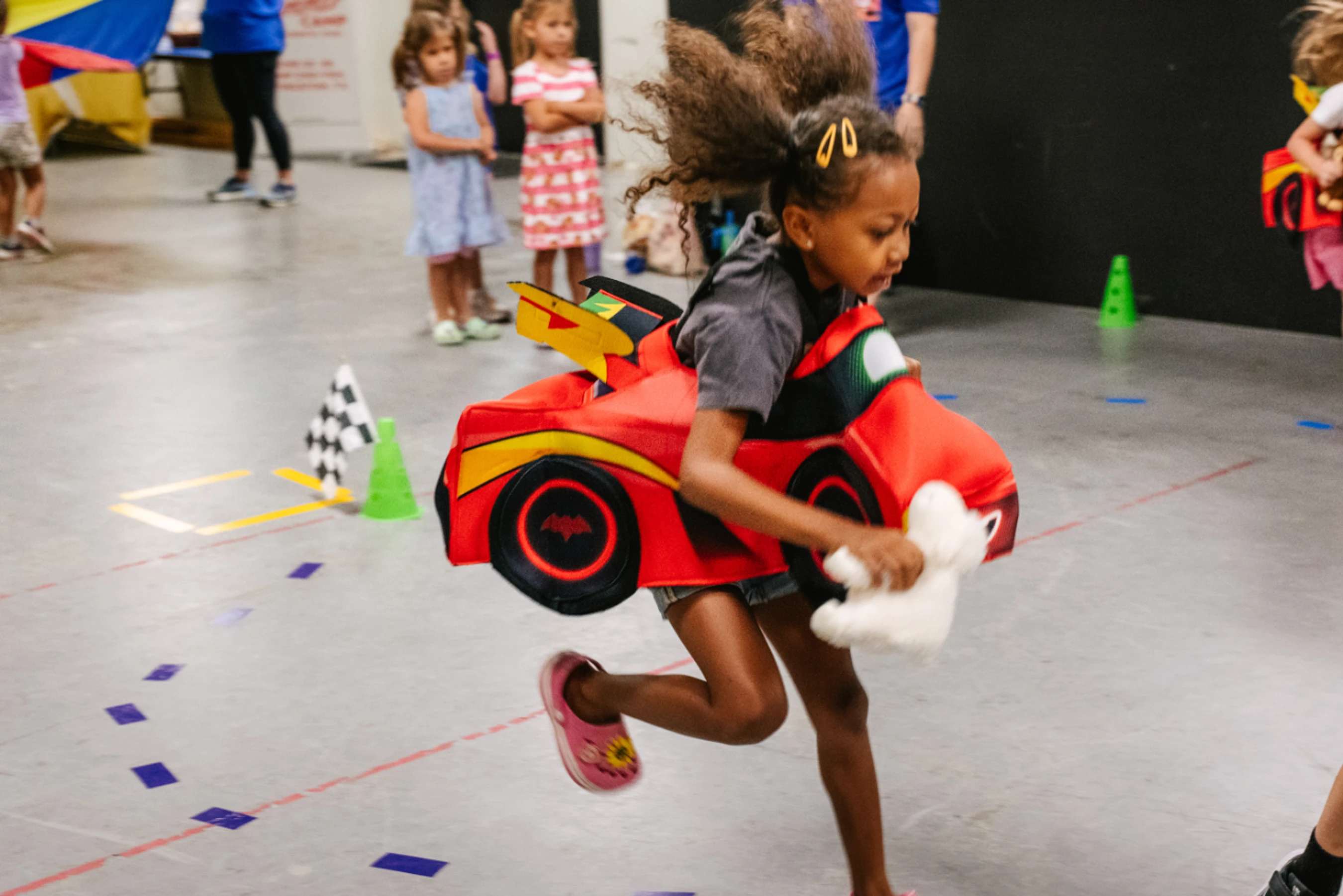 A young girl with curly hair runs with determination during an indoor race while wearing a red car costume and holding a stuffed animal.