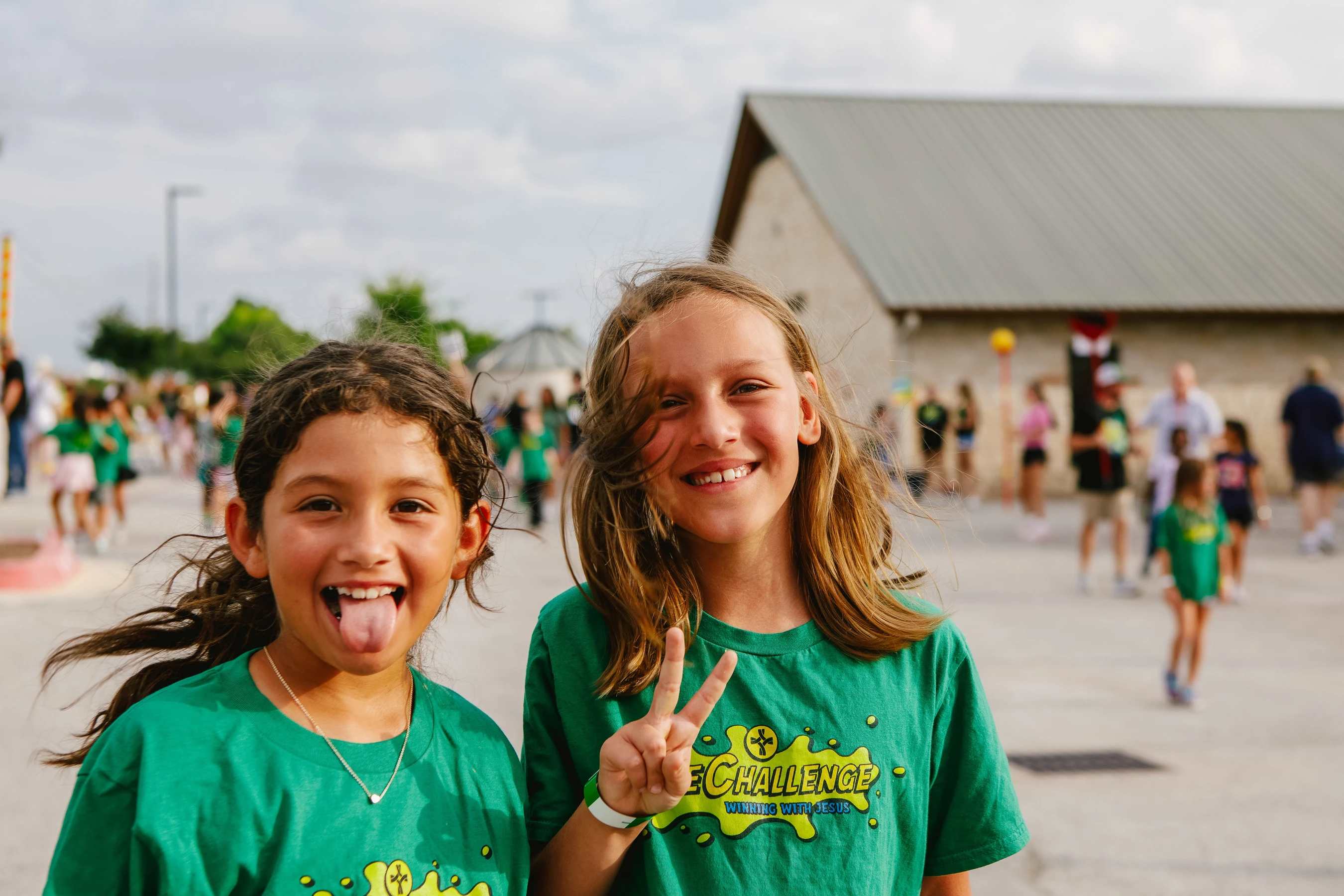 Two young girls in matching green t-shirts smile for the camera outdoors. One playfully sticks her tongue out while the other makes a peace sign.