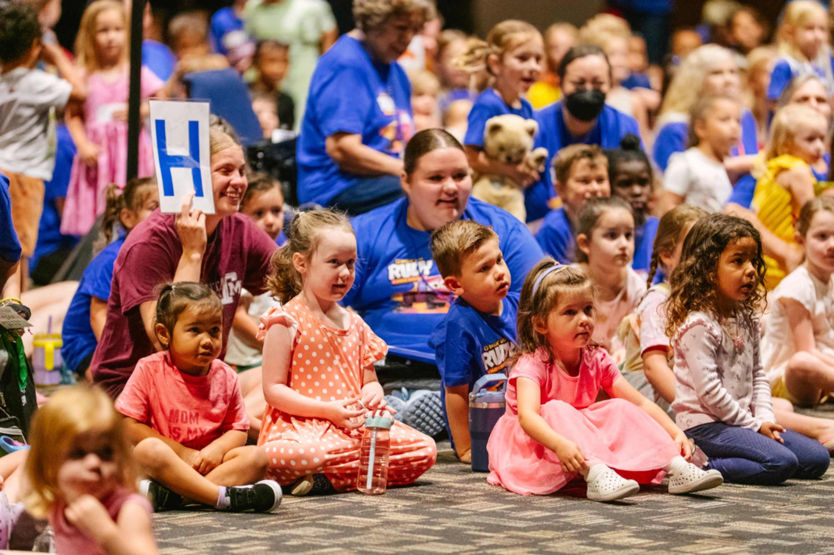 A large group of diverse preschoolers and toddlers sits on a carpeted floor, watching something off-camera with focused expressions during an event.