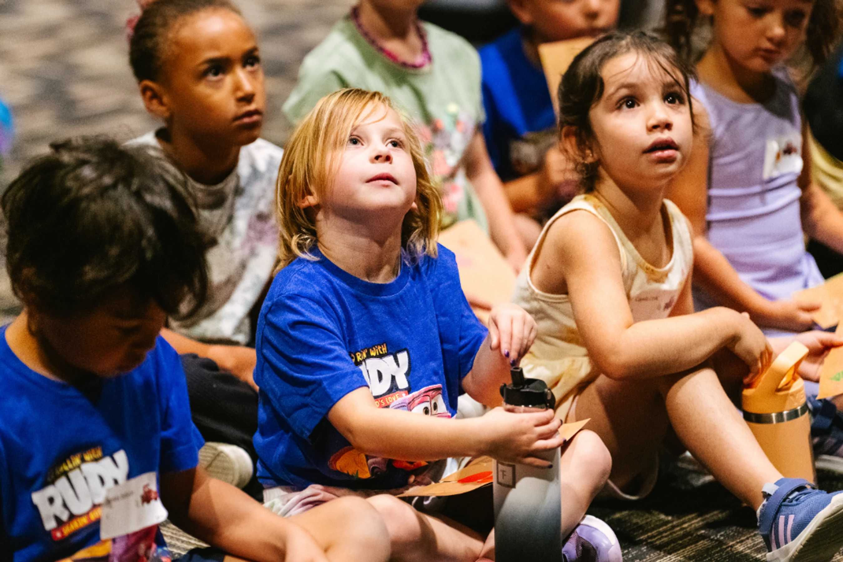 A diverse group of young children sit on a carpeted floor, looking up with rapt attention and curious expressions at something out of frame.