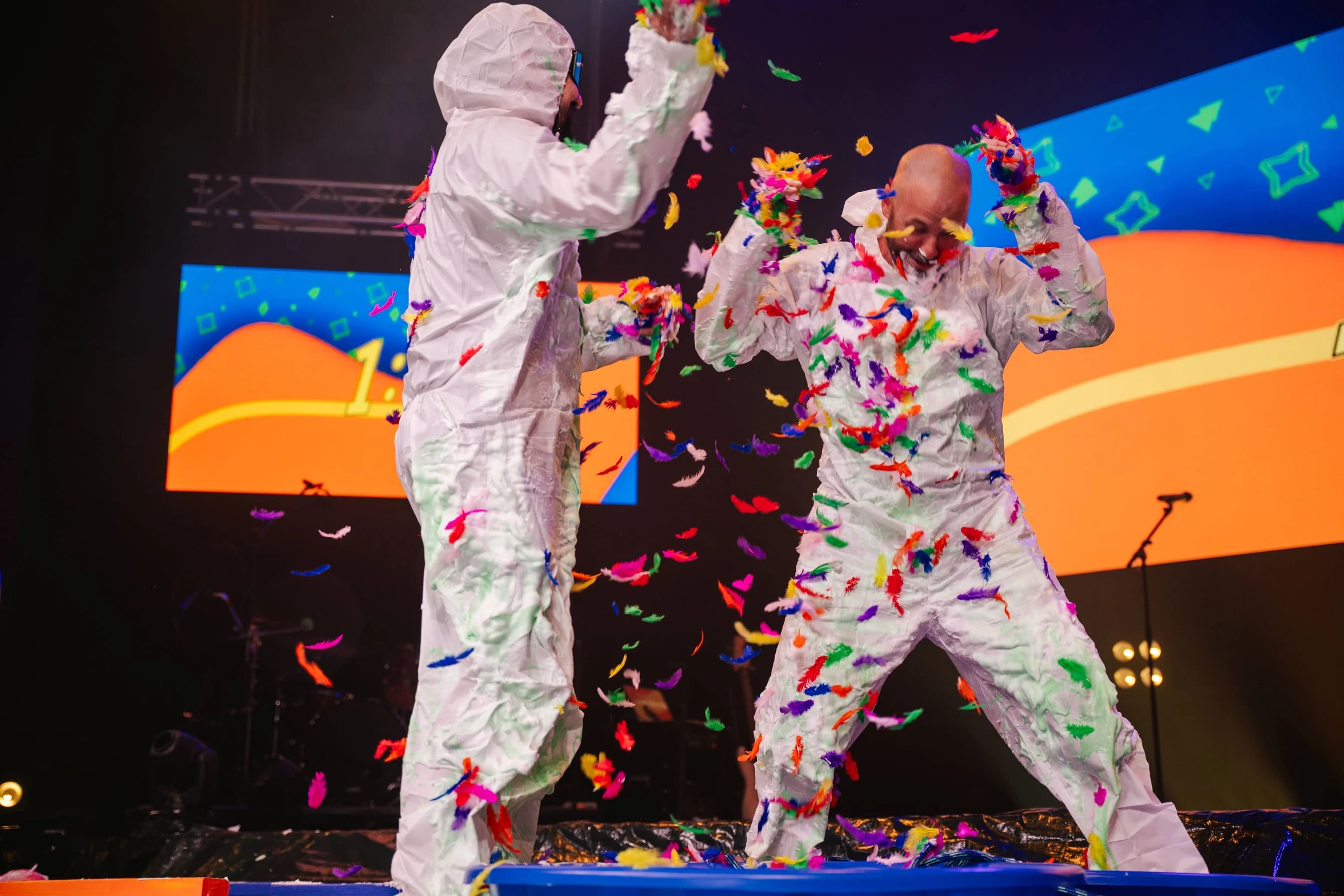 Two people in white protective suits laugh as they throw colorful feathers at each other on a stage, covered in foam and feathers.