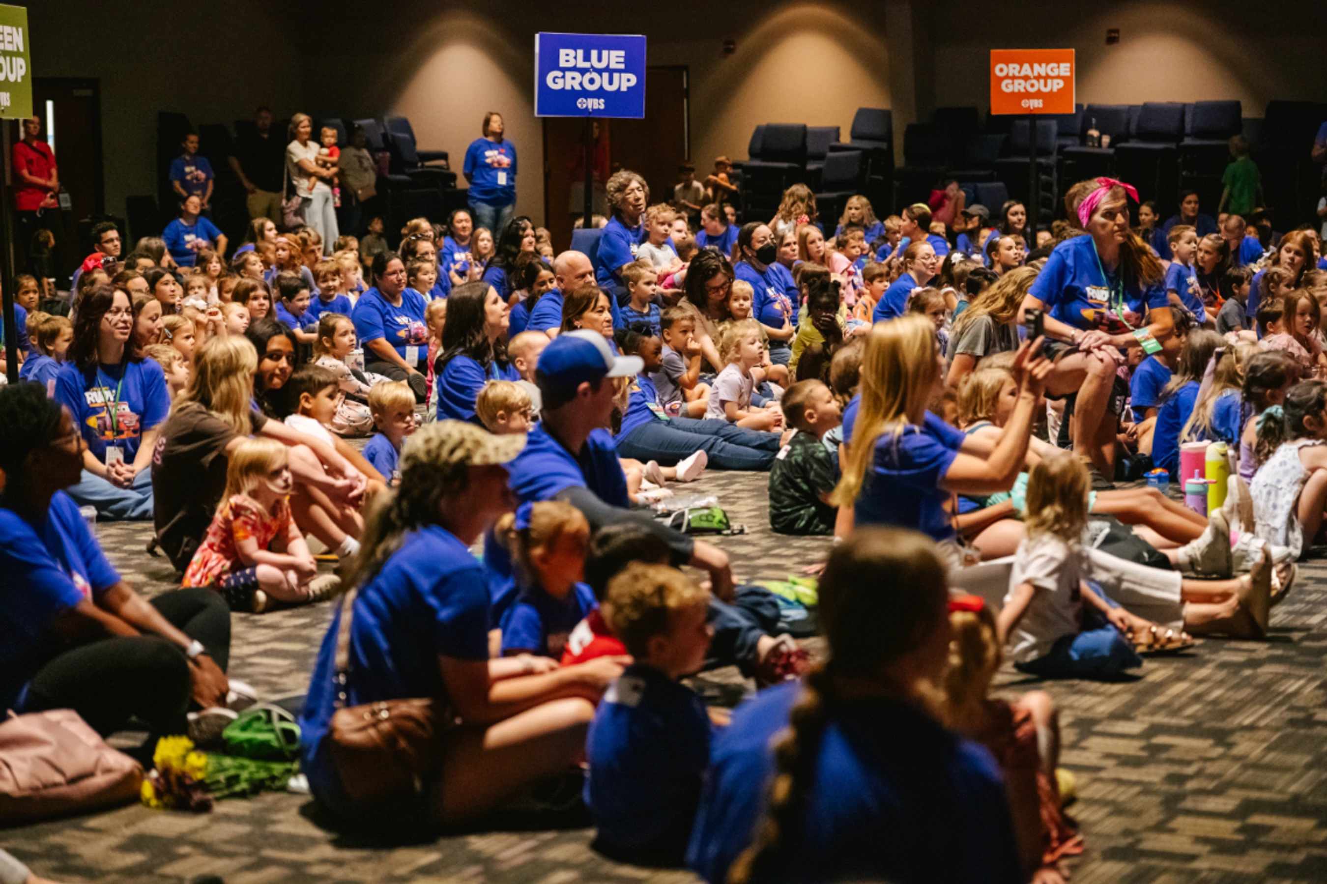 A large crowd of children and adult volunteers in matching blue shirts sit on the floor of an auditorium, watching a presentation during a VBS event.