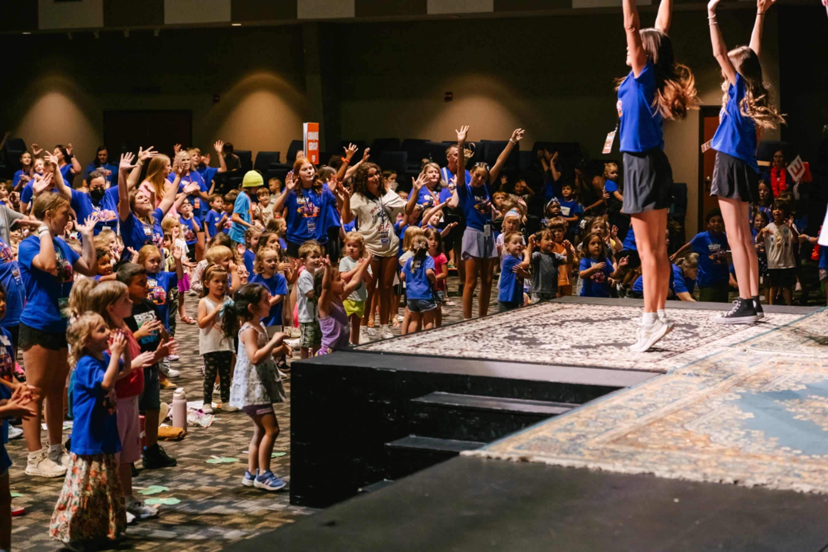 A large group of kids and counselors sing and dance in a hall. Two female leaders on a stage guide the crowd, who have their hands raised in excitement.