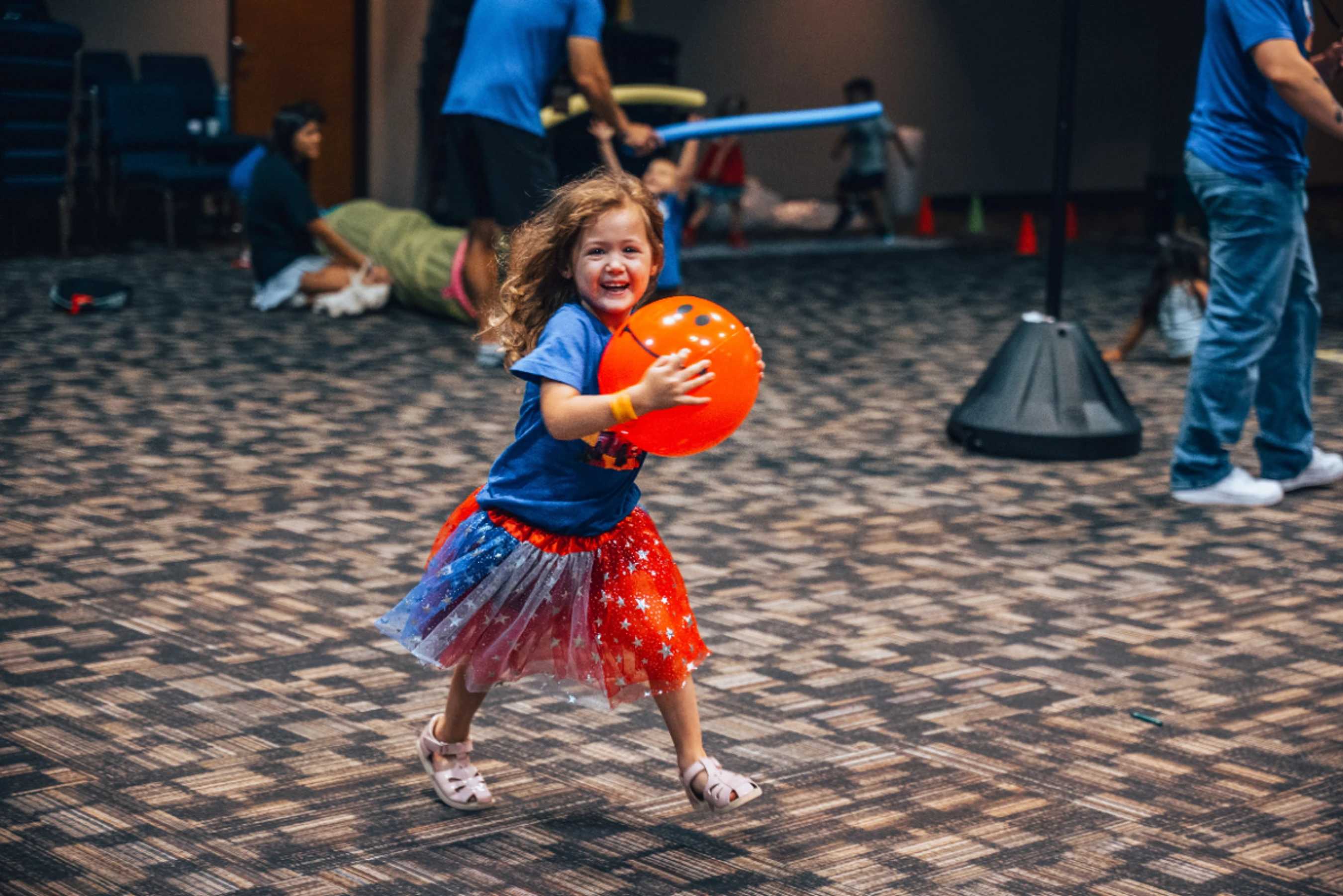 A young girl with curly hair smiles as she runs across a carpeted floor, holding a bright orange ball. She wears a blue t-shirt and a sparkly tutu.