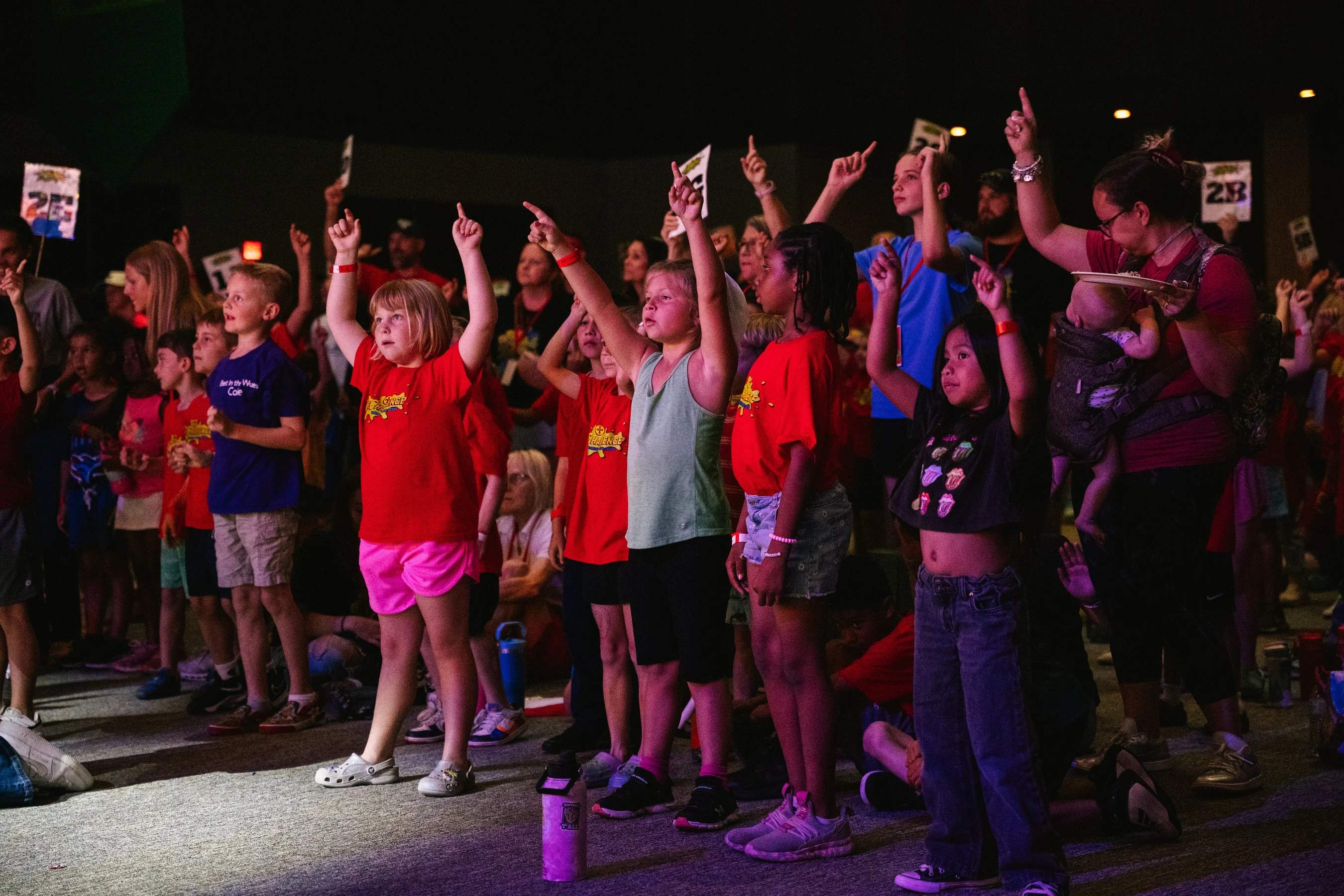 A diverse group of children and adults stand together in a dimly lit room, enthusiastically pointing their index fingers towards the ceiling.