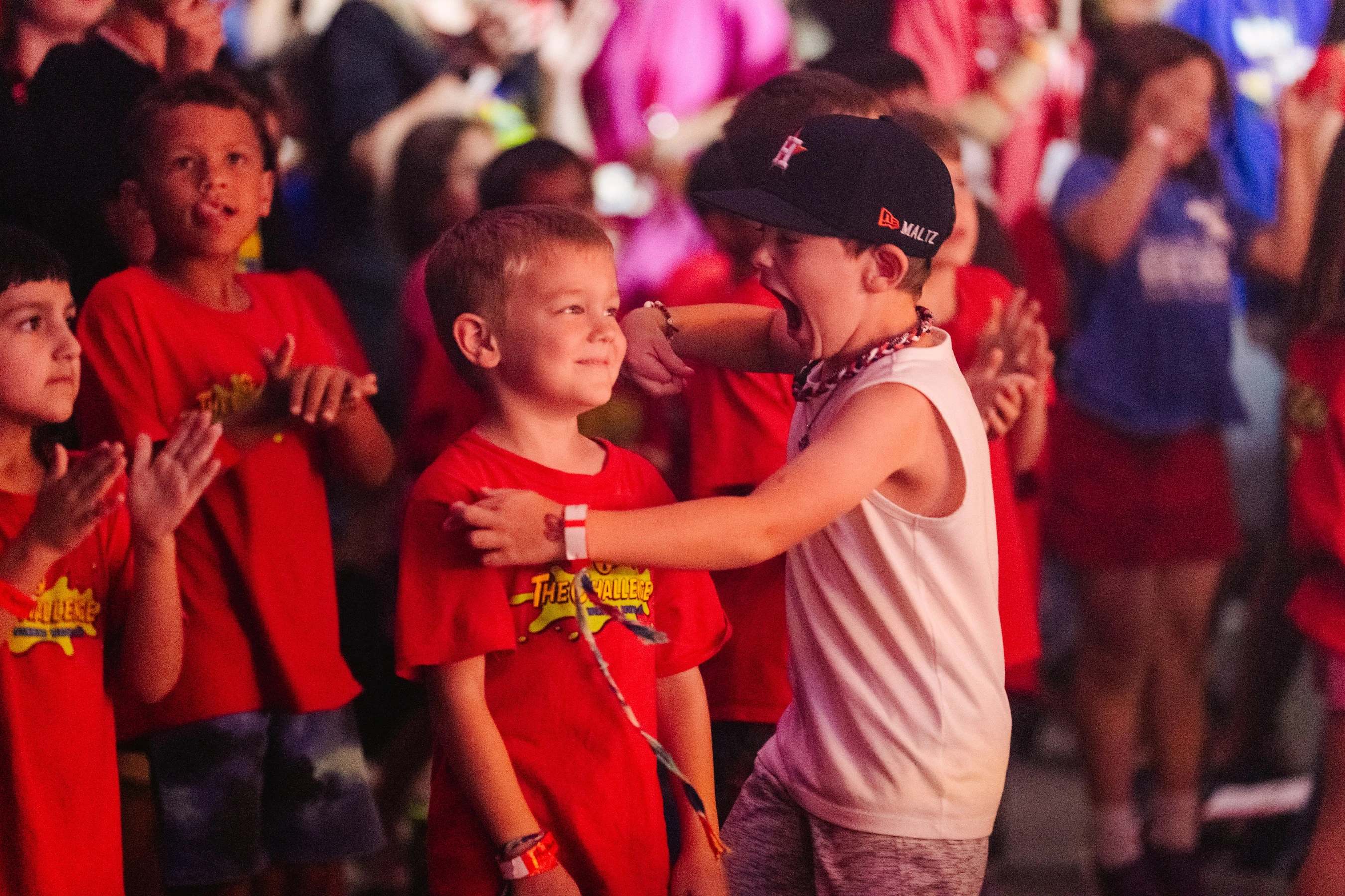 A young boy in a white tank top and baseball cap shouts with joy, his arms on the shoulders of another smiling boy in a red shirt amidst a crowd of children.