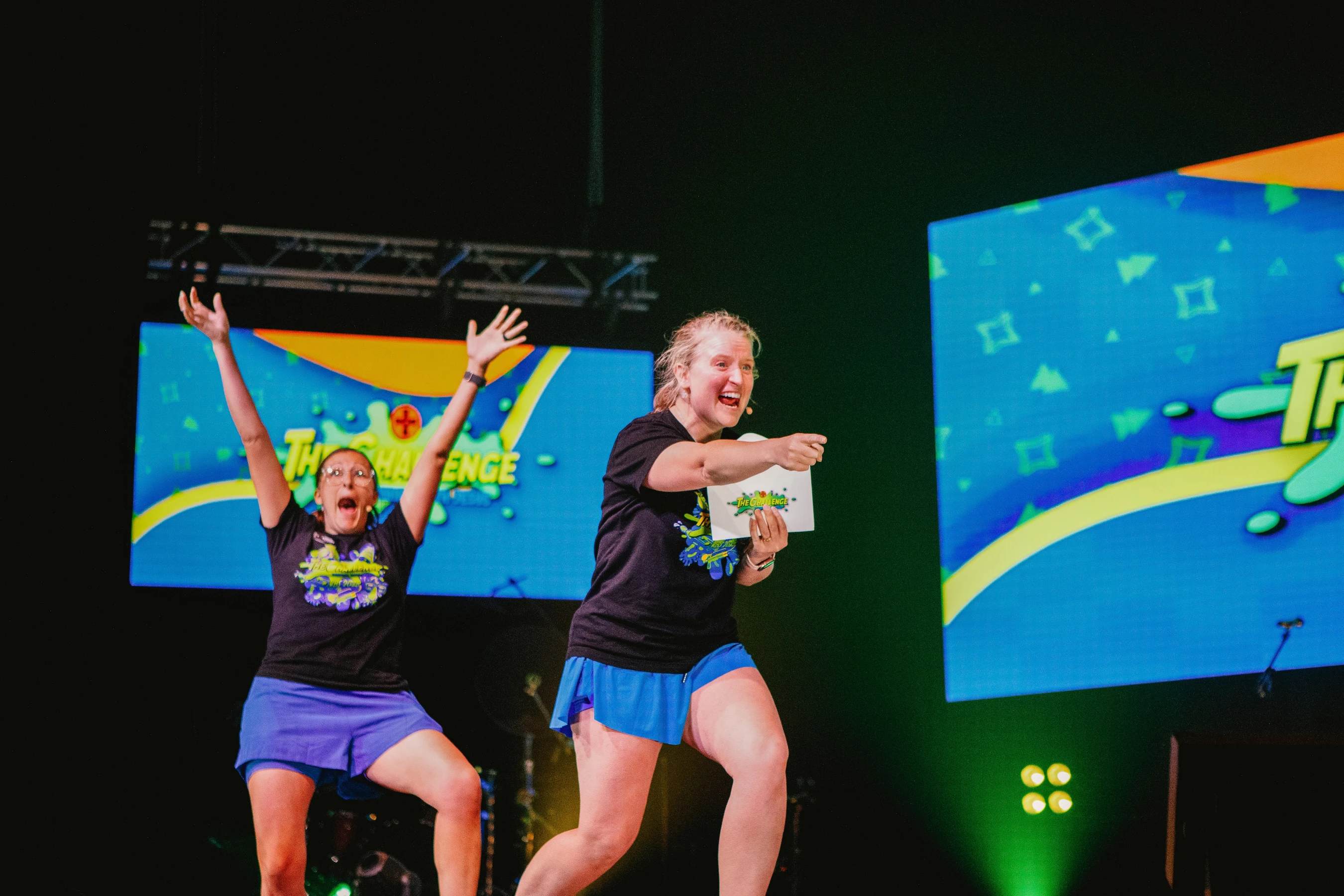 Two women on a brightly lit stage for an event. One woman raises her arms in celebration while the other smiles and points towards the audience.