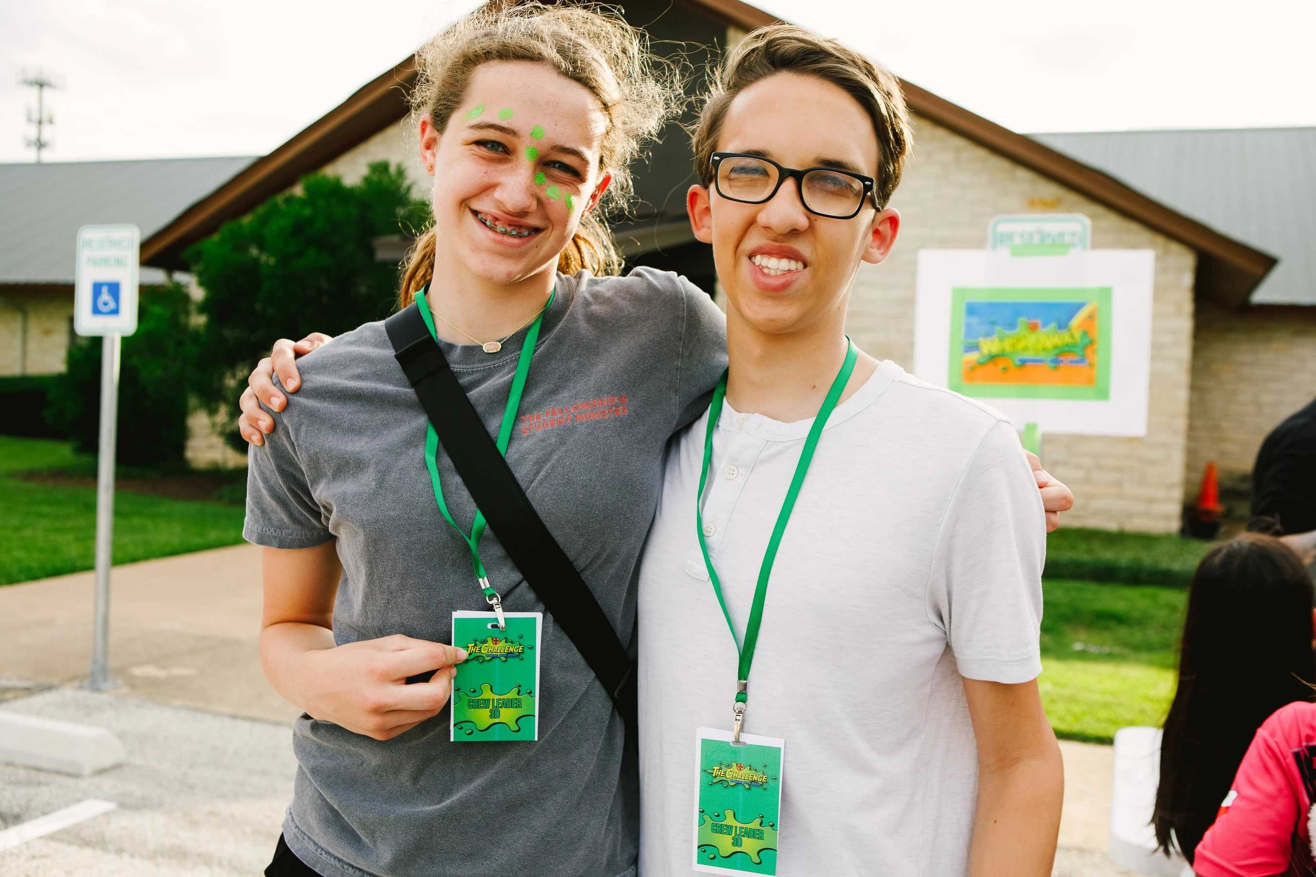 A teenage girl with green face paint and a boy with glasses smile for a photo. They have their arms around each other and wear 'Crew Leader' lanyards.