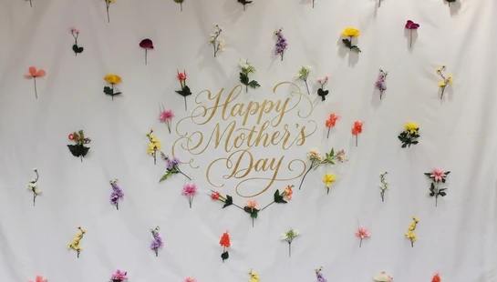A white fabric backdrop with 'Happy Mother's Day' in gold calligraphy, surrounded by a pattern of colorful artificial flowers.