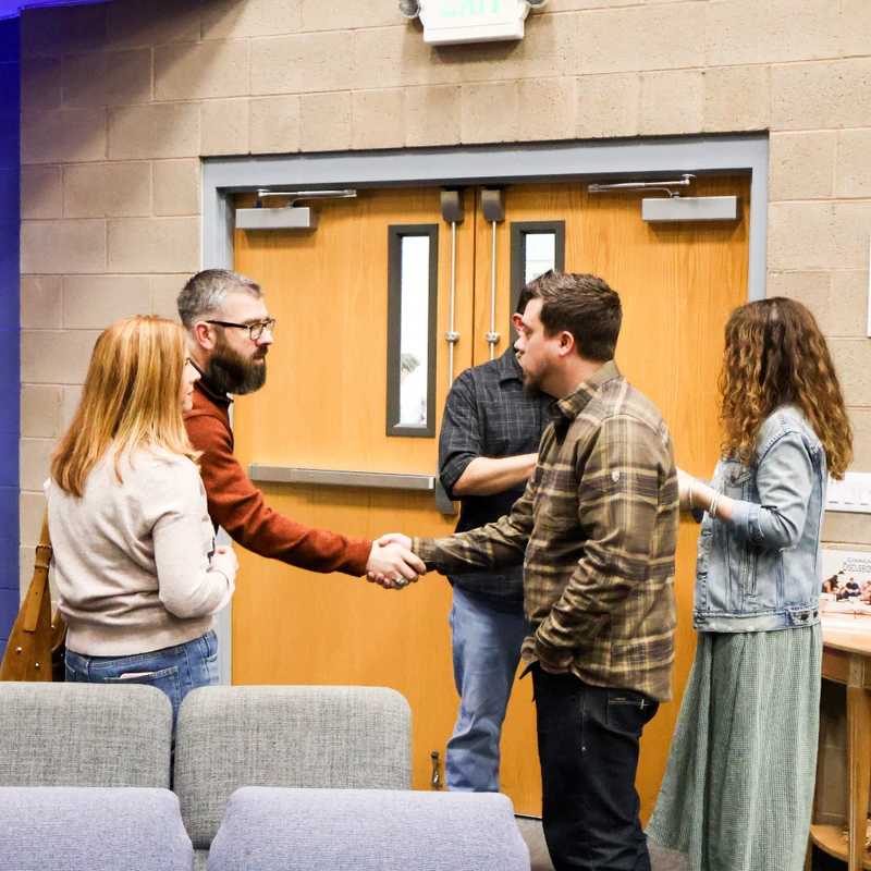 A group of adults stands in a church lobby, with two men shaking hands in the center. A young boy with headphones sits in a chair to the left.