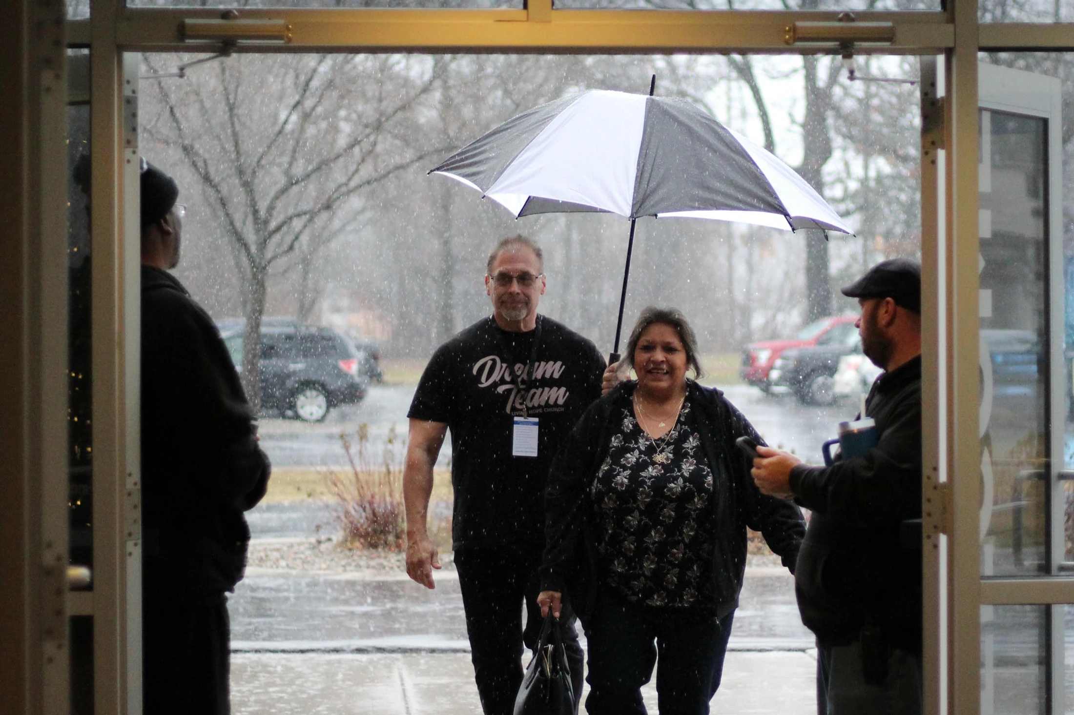 A man holds a black and white umbrella over a smiling woman as they walk in the rain towards a doorway, where they are greeted by two other men.
