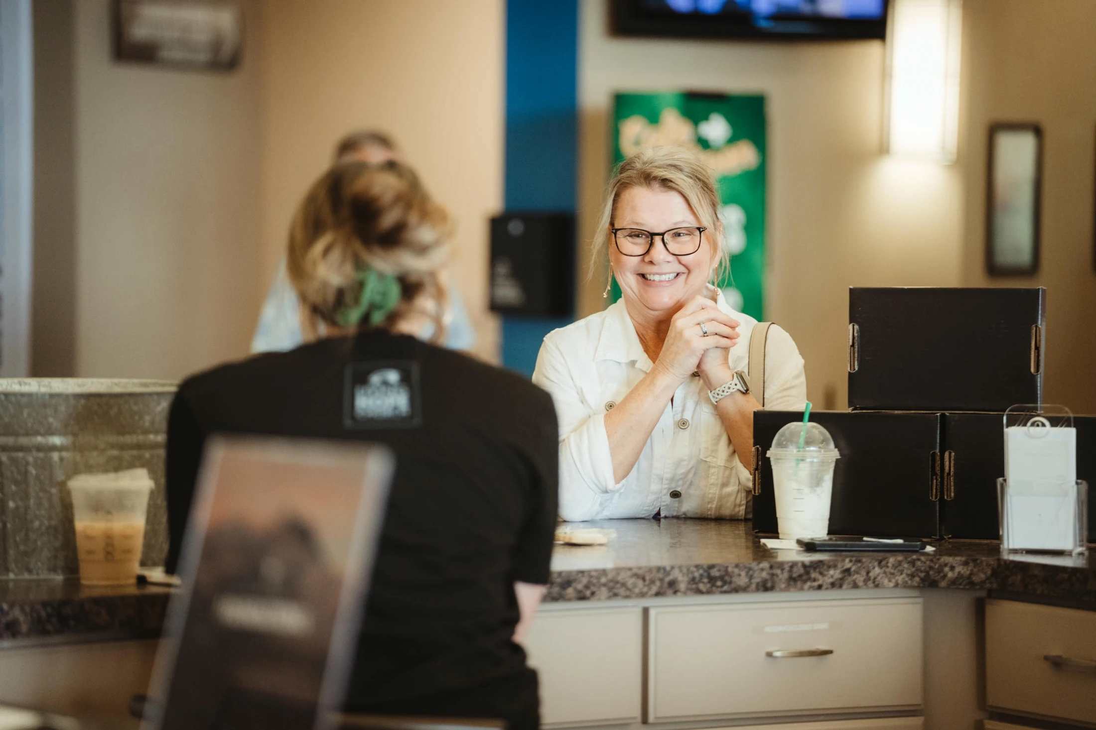 A smiling blonde woman with glasses stands at a counter, engaging in a friendly conversation with a person whose back is to the camera.