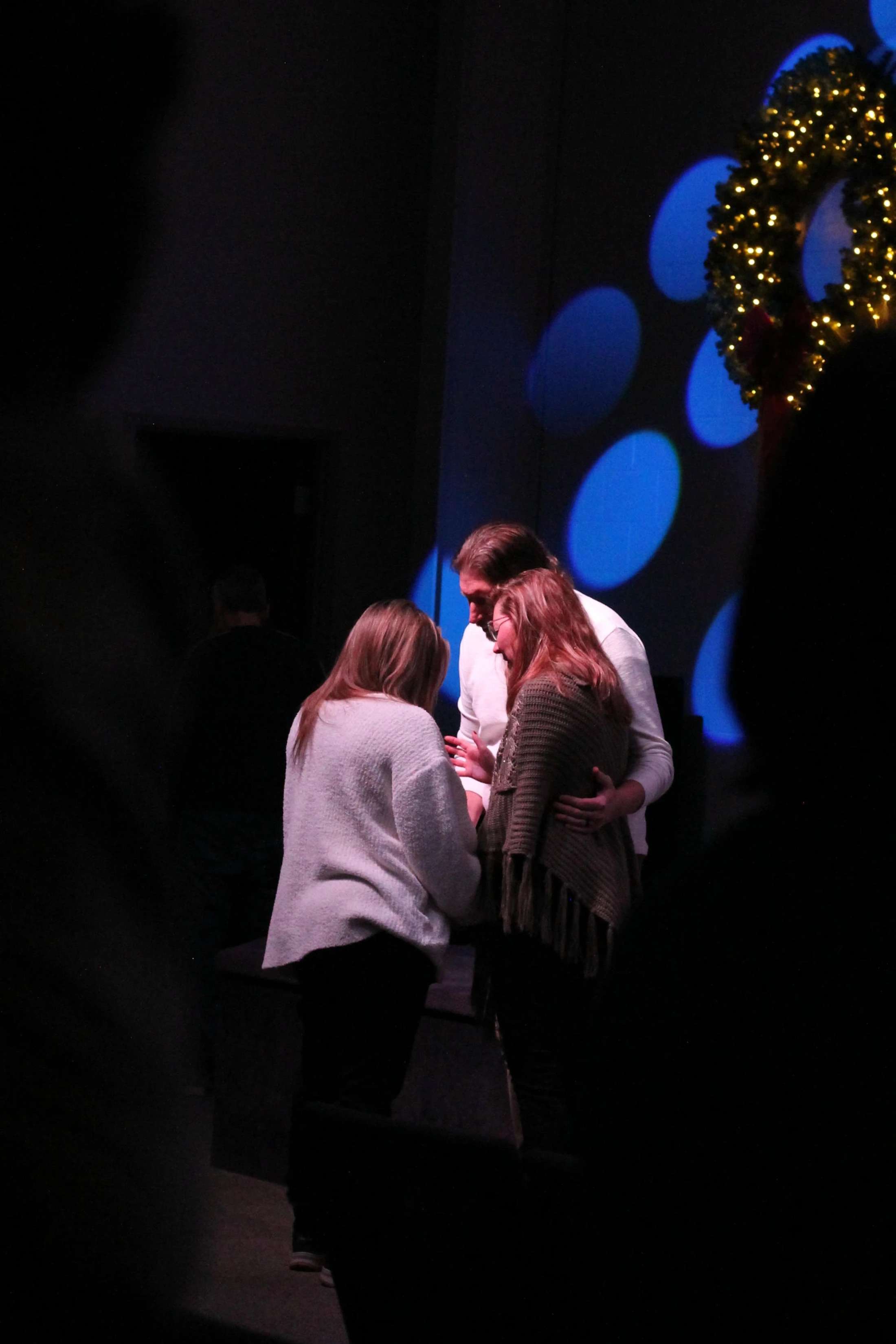 Three people stand huddled together in a spotlight on a dark stage, seemingly in prayer, with a lit Christmas wreath in the background.