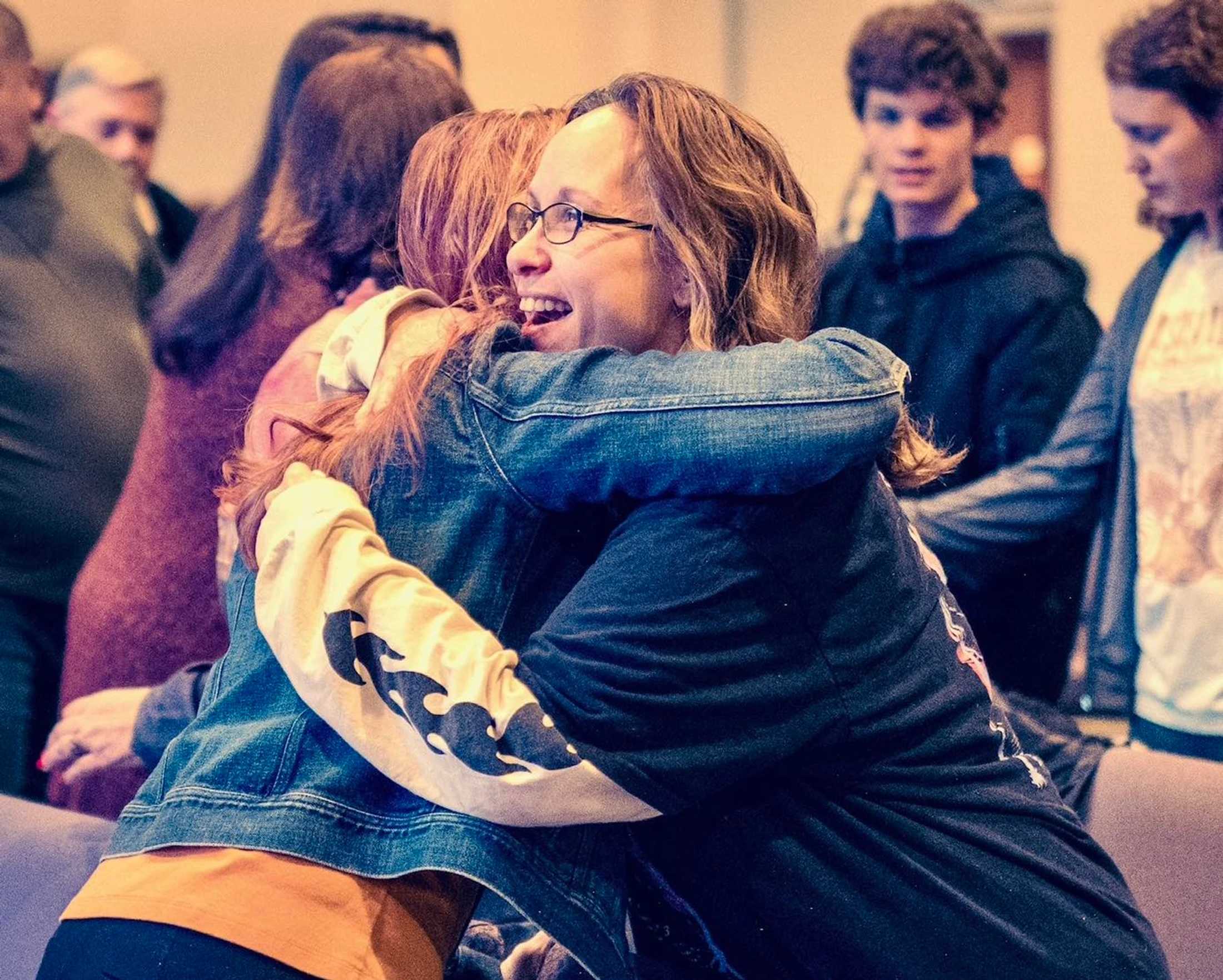 Two women share a warm, smiling hug in a room full of people. The woman in focus wears glasses and a dark shirt, embracing a friend in a denim jacket.