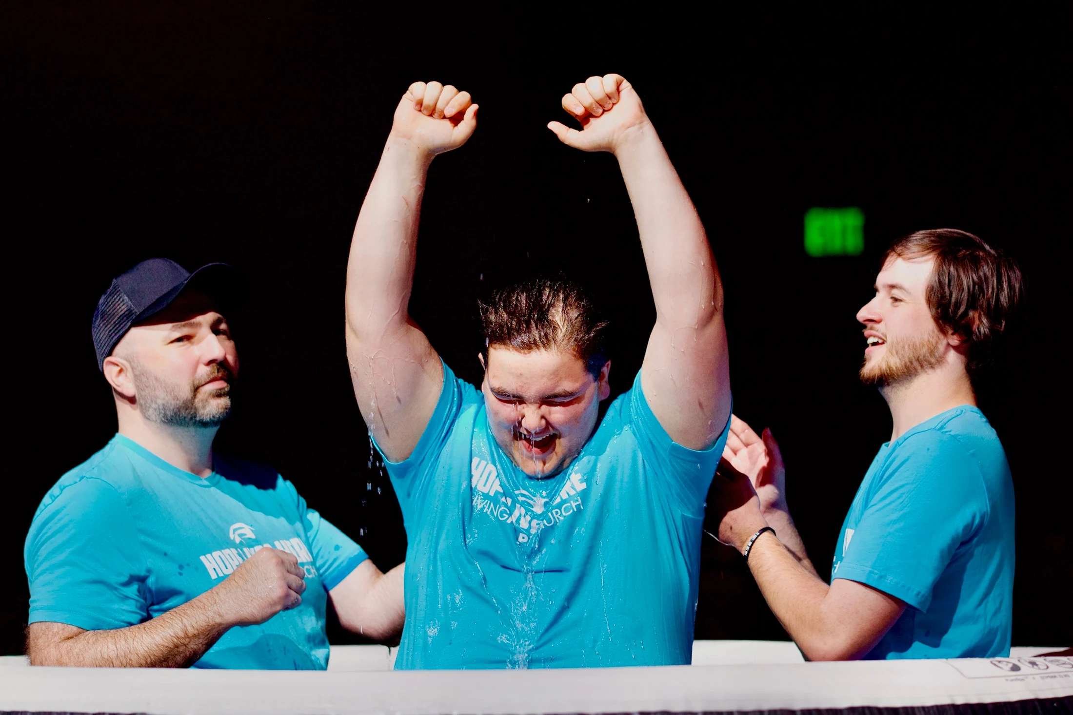 A young man raises his arms in celebration as he emerges from the water during a baptism, flanked by two other men in a baptismal pool.