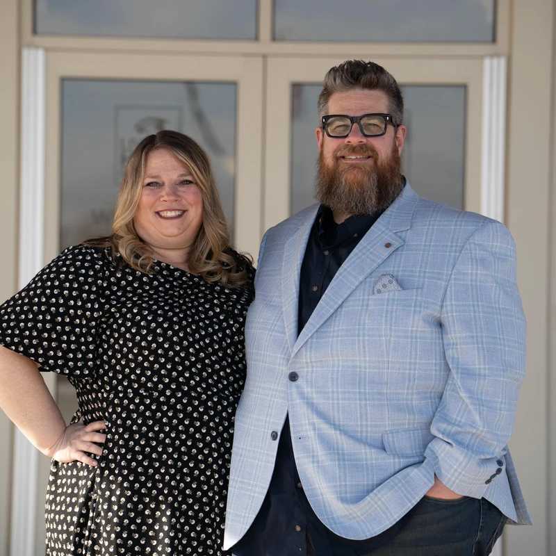 A smiling man and woman pose for a photo in front of glass doors. The woman wears a patterned dress, and the man wears a light blue plaid jacket and glasses.