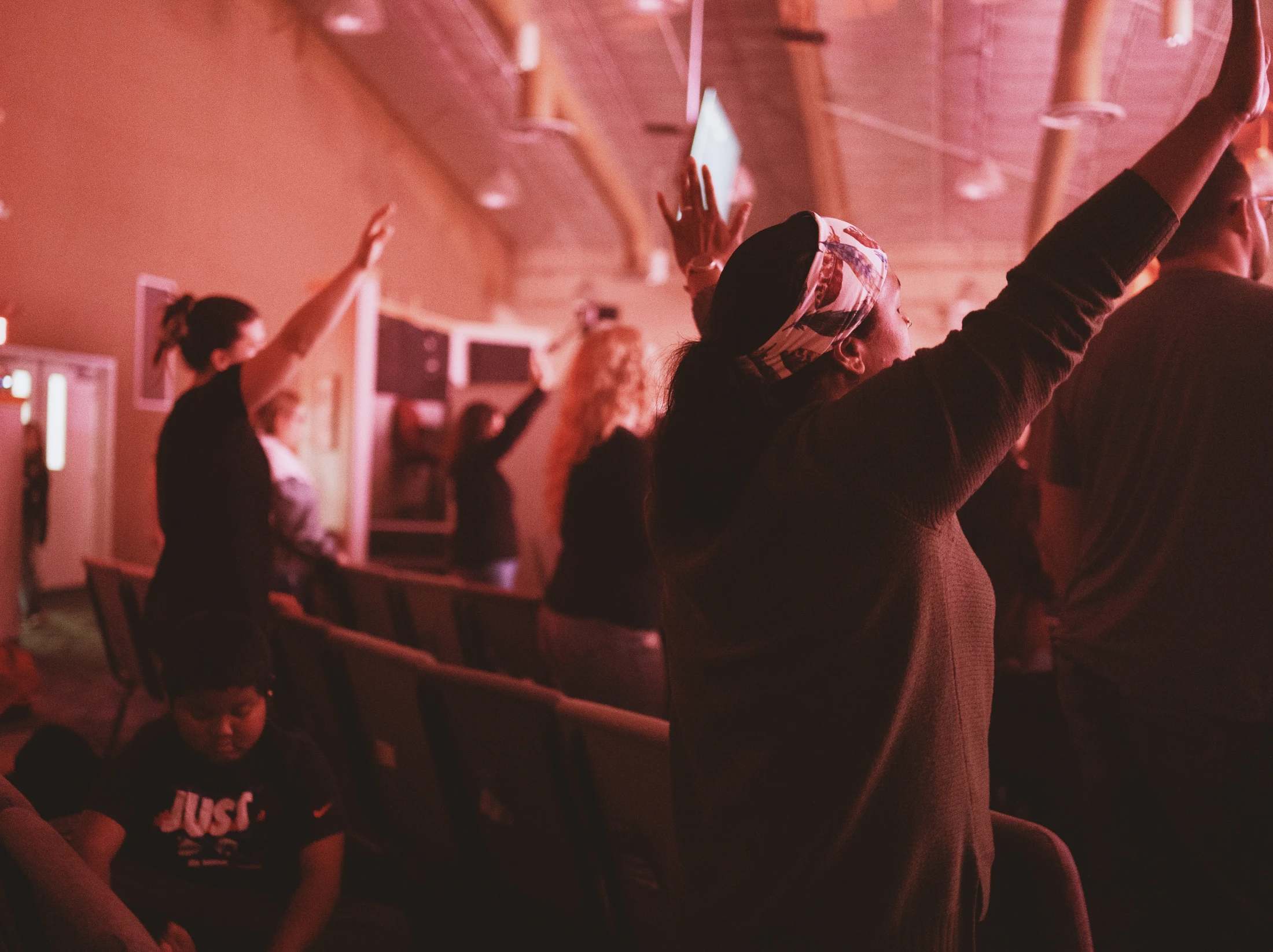 People in a dimly lit room with a red hue have their hands raised in worship. The focus is on a woman from behind, with others visible in the background.