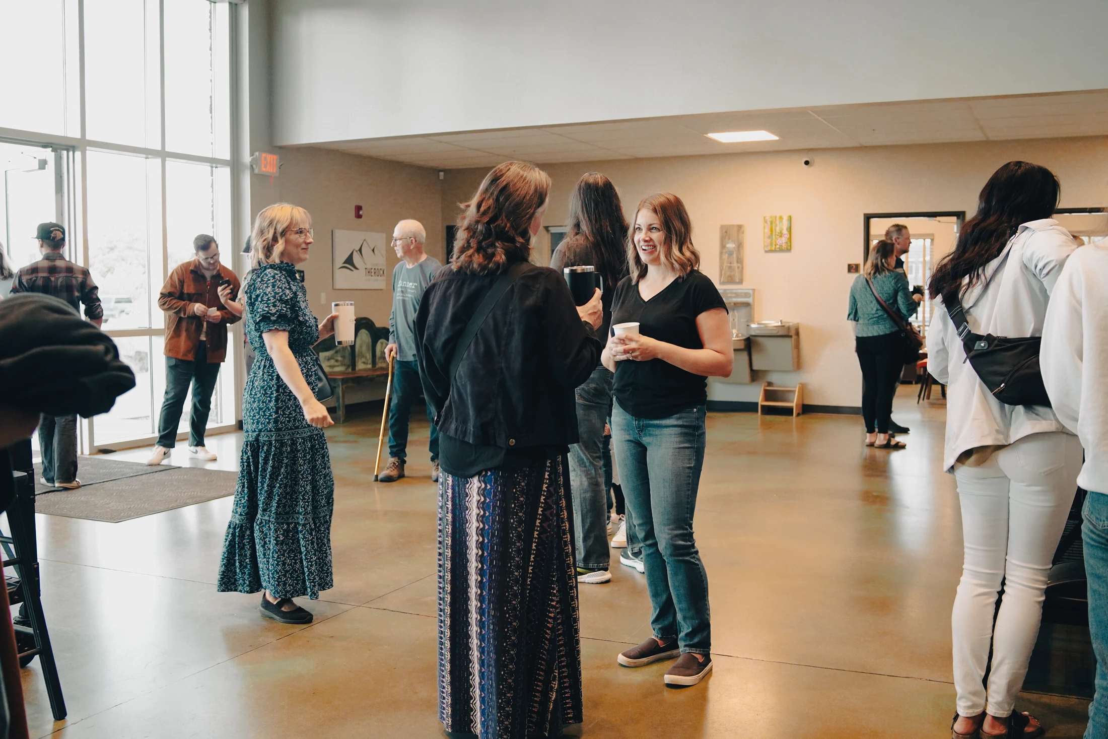 A group of people stand in a brightly lit lobby, talking in small groups and holding cups. Three women are in conversation in the center of the room.