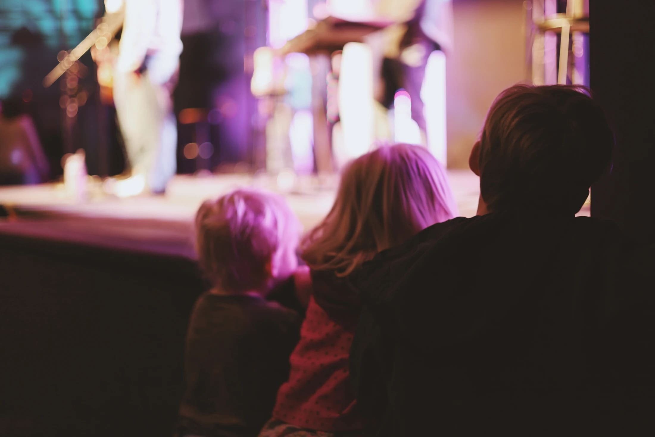 From behind, a parent and two small children watch a band perform on a stage illuminated by purple and pink lights.