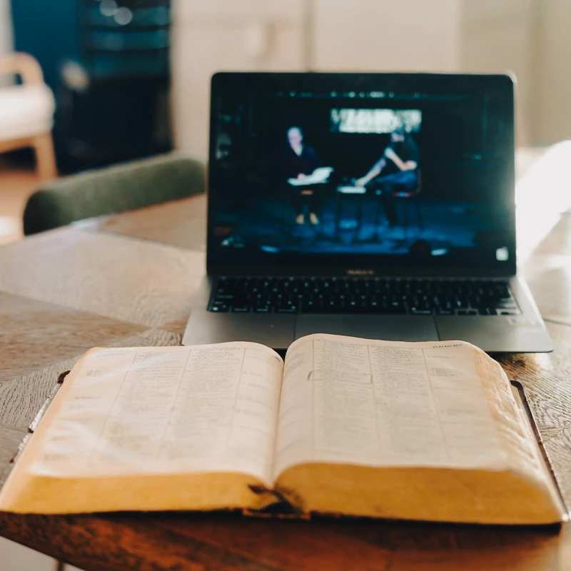 An open Bible rests on a wooden table in front of a laptop streaming what appears to be an online church service or sermon in a warmly lit room.