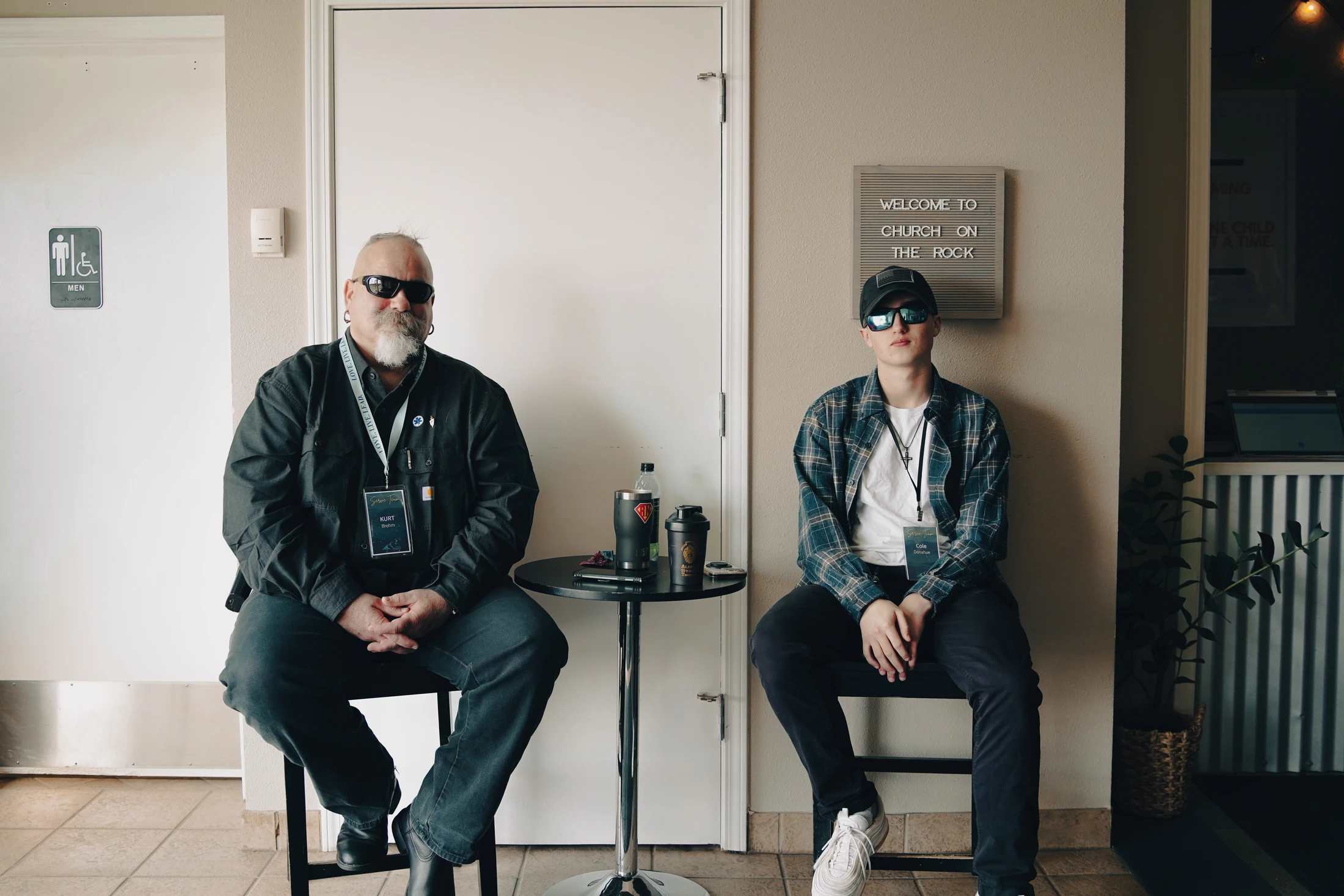 An older man and a younger man, both wearing sunglasses and serve team lanyards, sit on stools in a church hallway next to a 'Welcome' sign.