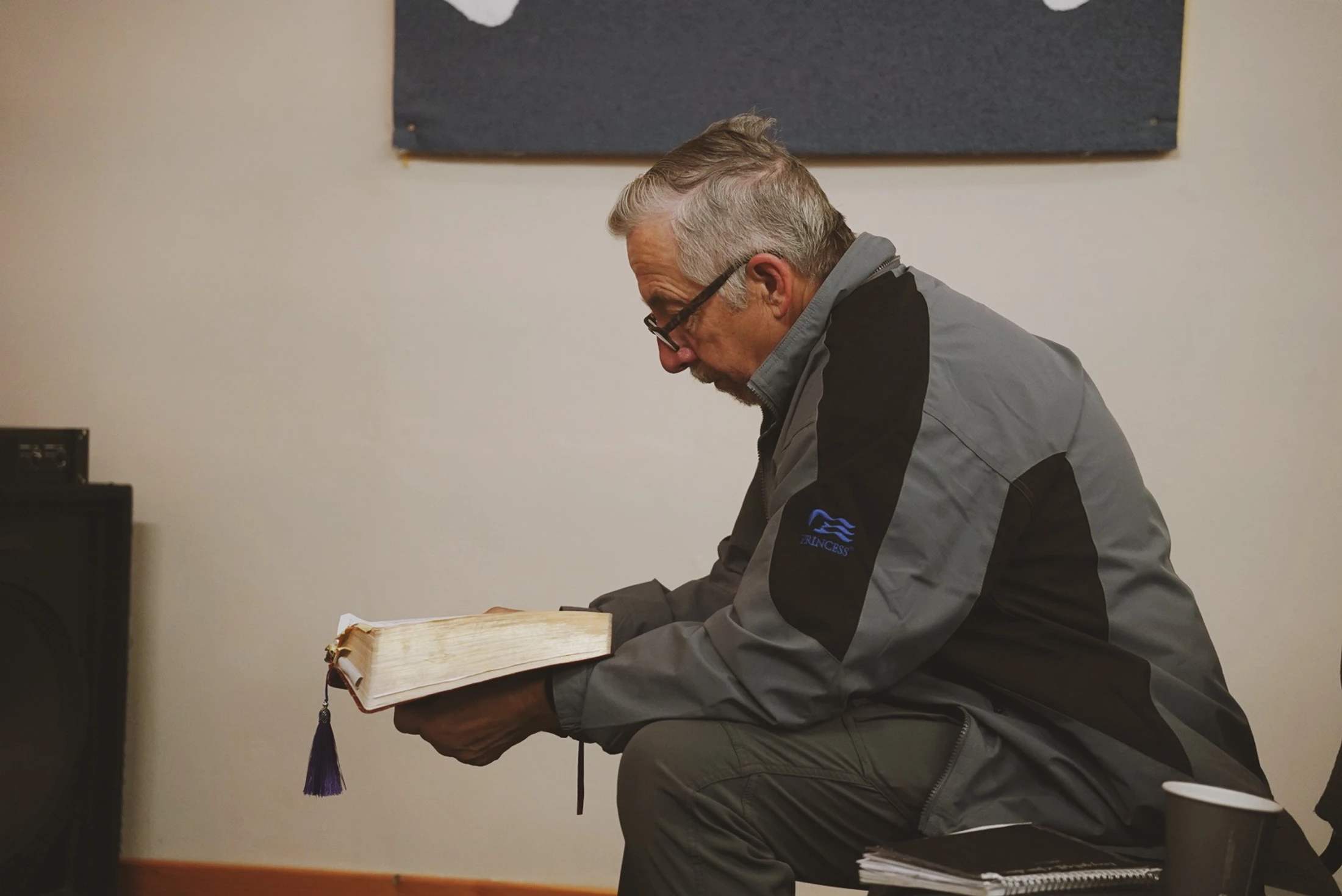 Side profile of an older man with gray hair and glasses, sitting down and attentively reading a large, thick book with a purple tassel bookmark.