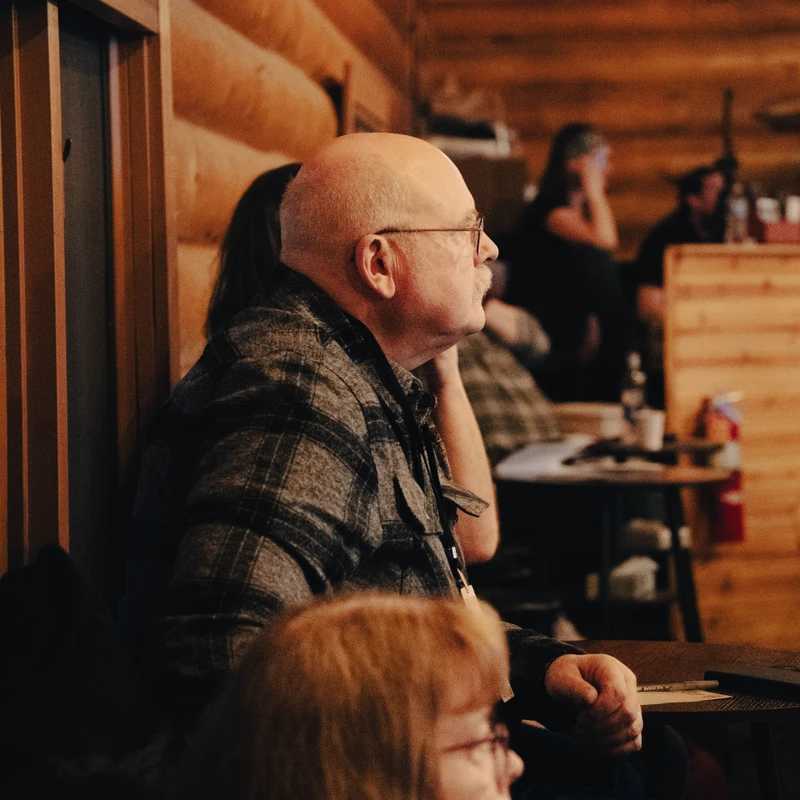 A bald man with glasses and a mustache, wearing a plaid shirt, sits in profile and listens intently inside a warmly lit room with wooden log walls.
