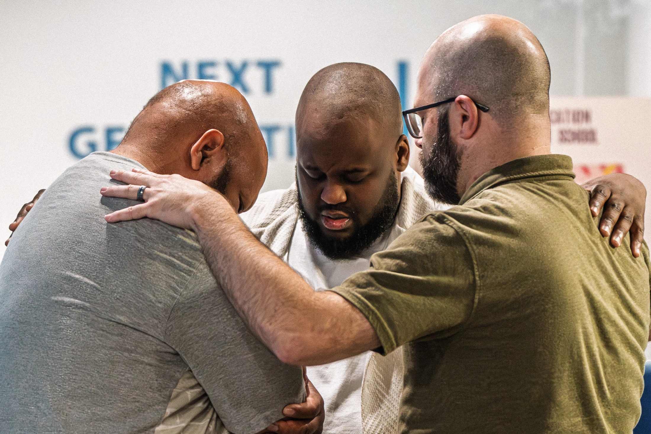 Three diverse men are huddled together with their arms around each other, heads bowed and eyes closed in a moment of emotional prayer.