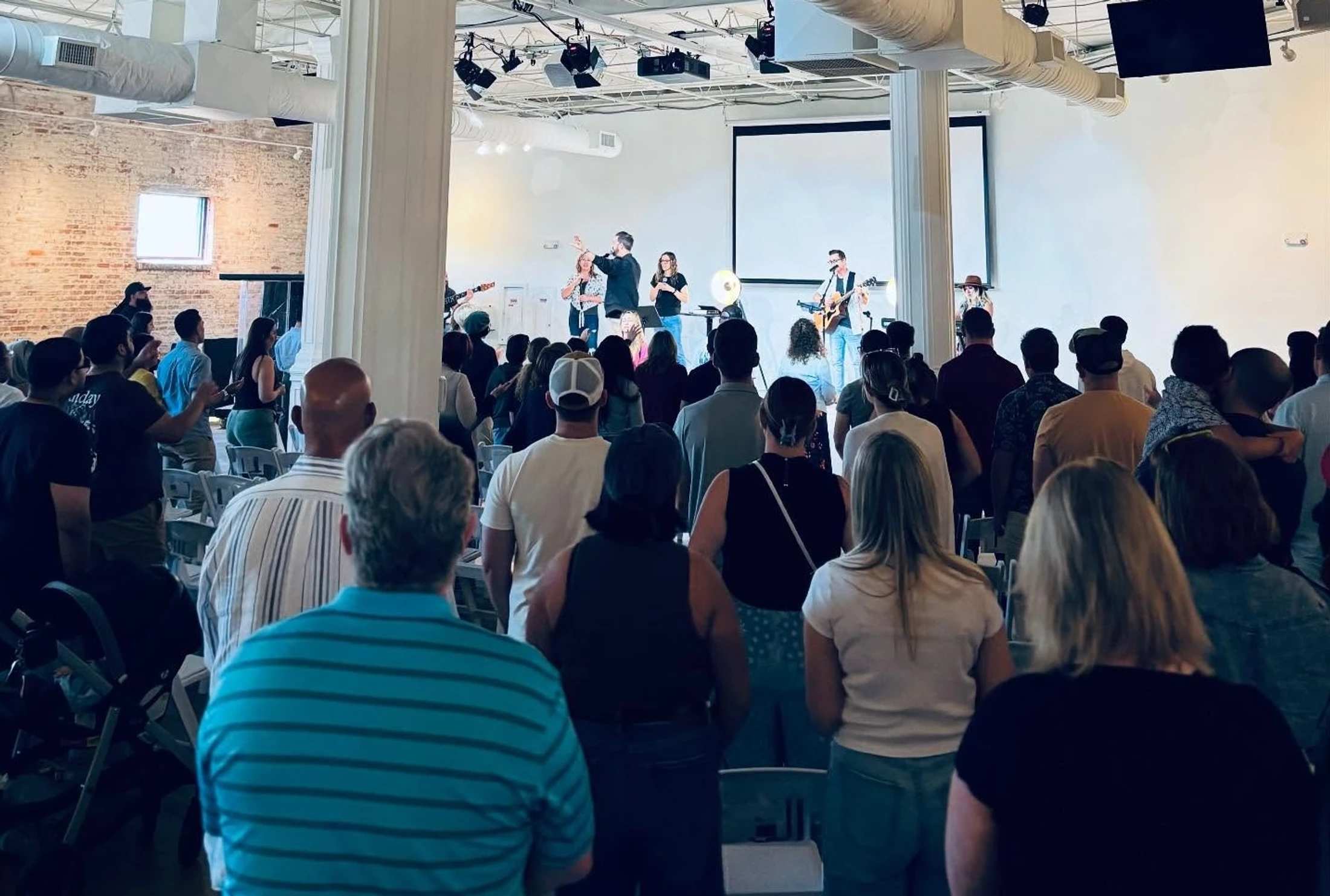 From behind, a diverse congregation stands and watches a band perform on a well-lit stage in a modern, spacious room with white pillars and exposed brick.