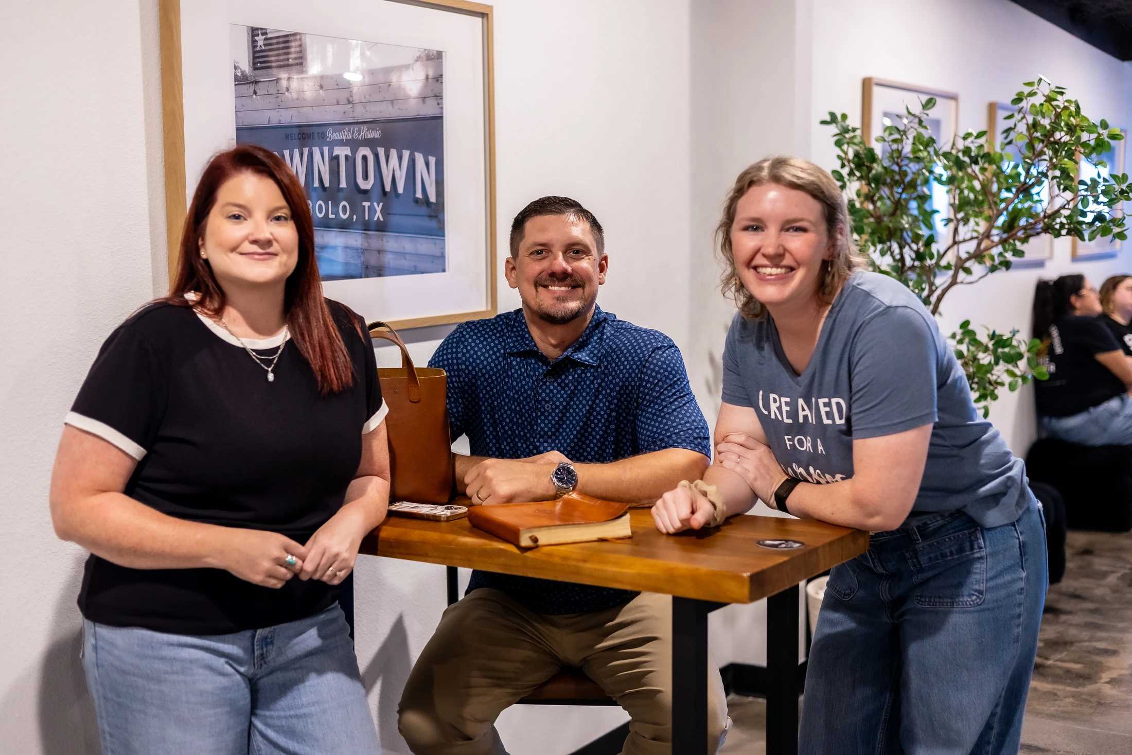 A man sits smiling between two smiling women who are standing at a tall wooden table. They are posing for a photo in a casual indoor setting.