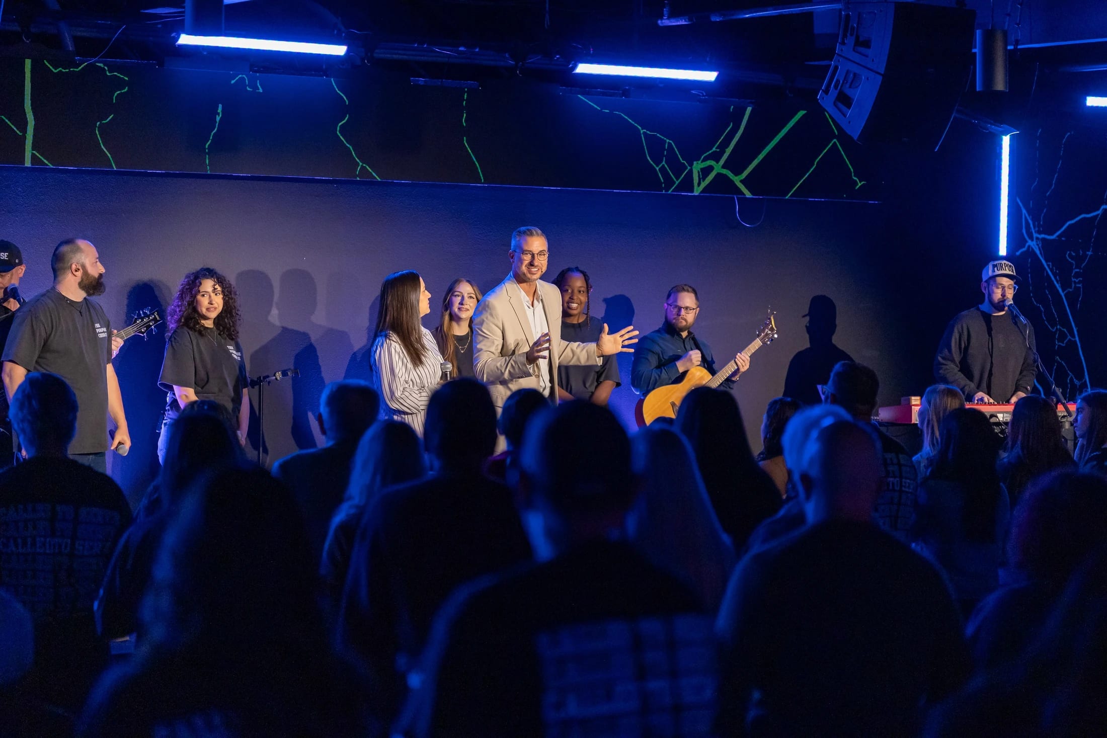 A man in a tan suit speaks on a stage with a band, addressing an audience in a dimly lit room with blue lighting and abstract green line art on the wall.