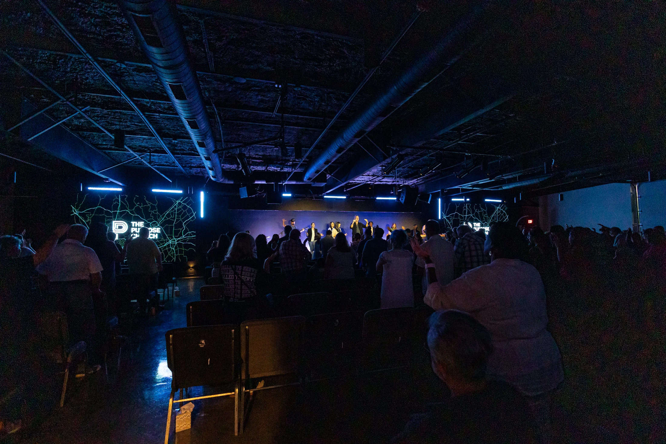 A wide shot from the back of a dimly lit room showing a congregation standing and worshipping. A band performs on a stage lit with cool blue lights.