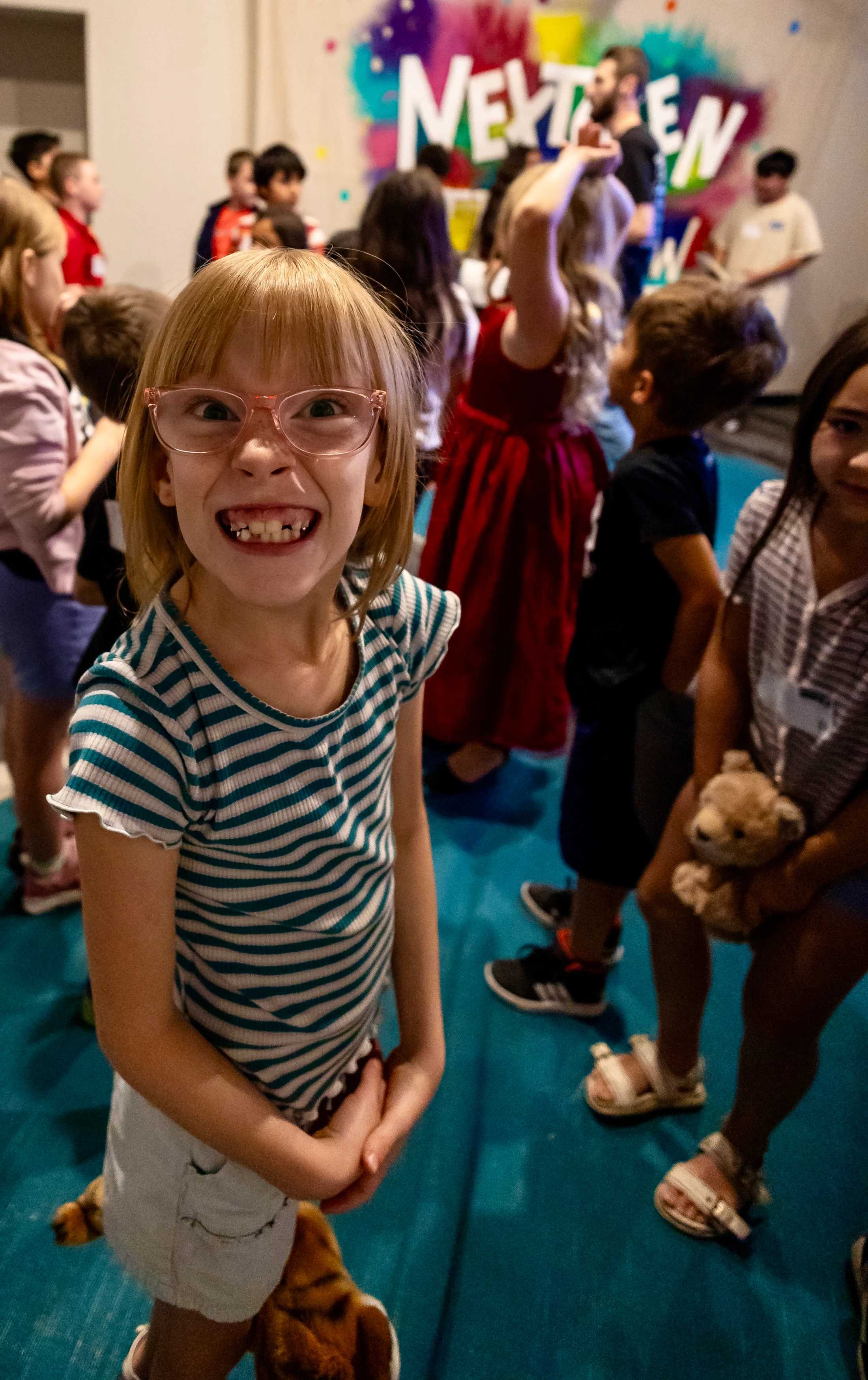 A young girl with blonde hair and pink glasses stands in a crowd, smiling widely at the camera and showing her teeth in a playful, silly grin.