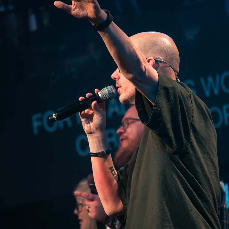 A bald man on a dimly lit stage sings into a microphone. He wears a dark t-shirt and has one hand raised in a gesture of worship or praise.