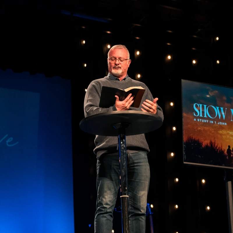 A middle-aged man with glasses stands on a stage behind a lectern, reading from a book. A screen next to him reads "A STUDY IN 1 JOHN."