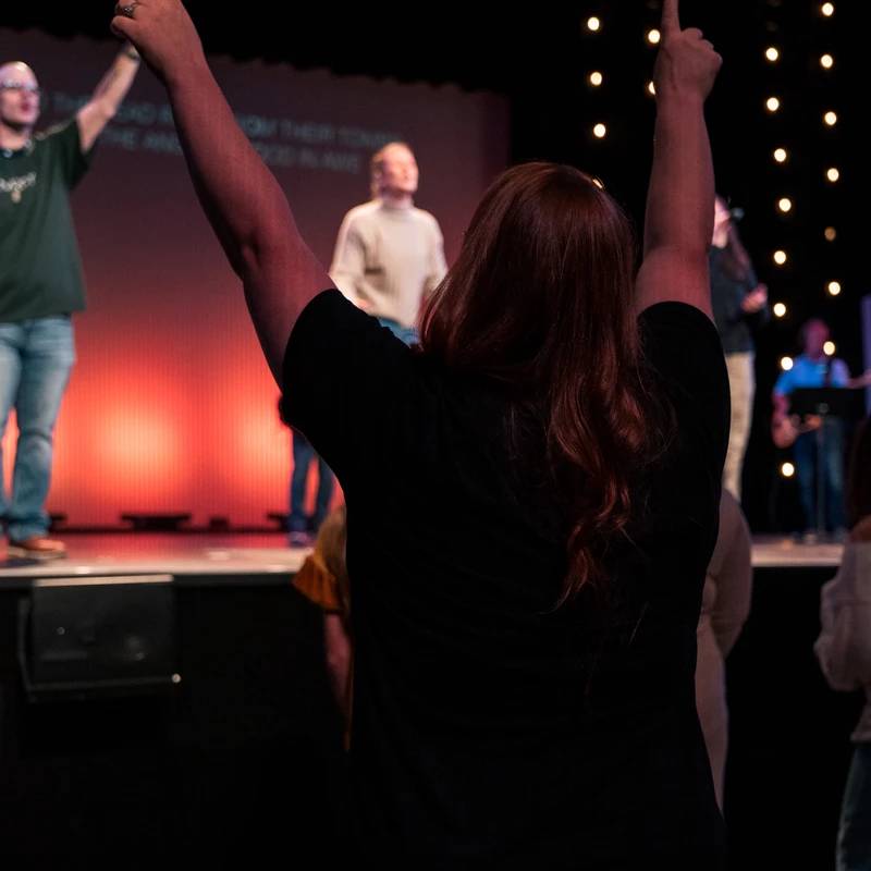 A woman with red hair, seen from behind, raises her arms in worship during a church service. A band is visible on the dimly lit stage in front of her.