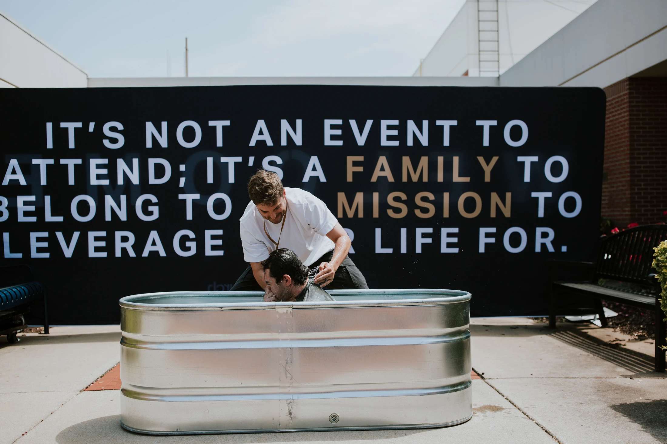 A pastor baptizes a man by immersion in a metal stock tank outdoors. Behind them is a large black banner with white and gold text about community.