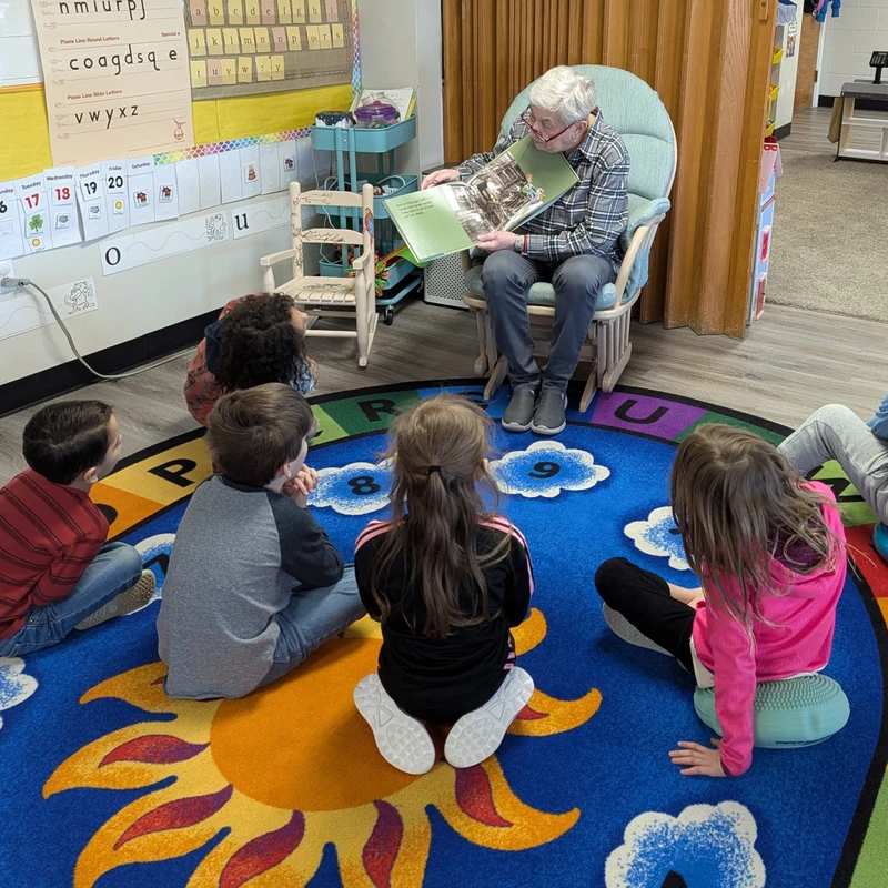 An elderly woman sits in a rocking chair reading a picture book to a small group of young children sitting on a colorful alphabet rug in a classroom.