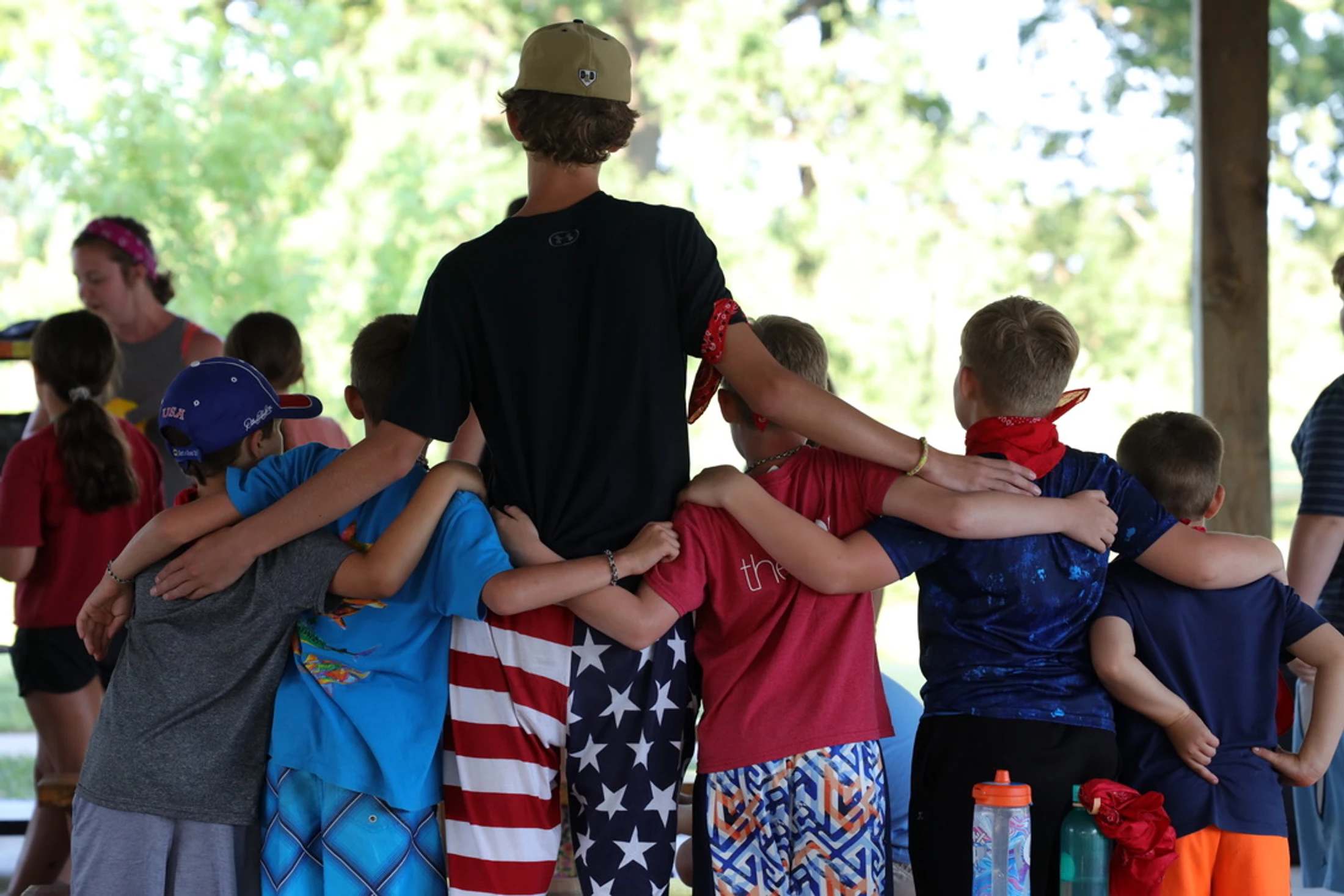 A group of young campers and a counselor seen from behind, huddled together with their arms around each other's shoulders in a show of unity.