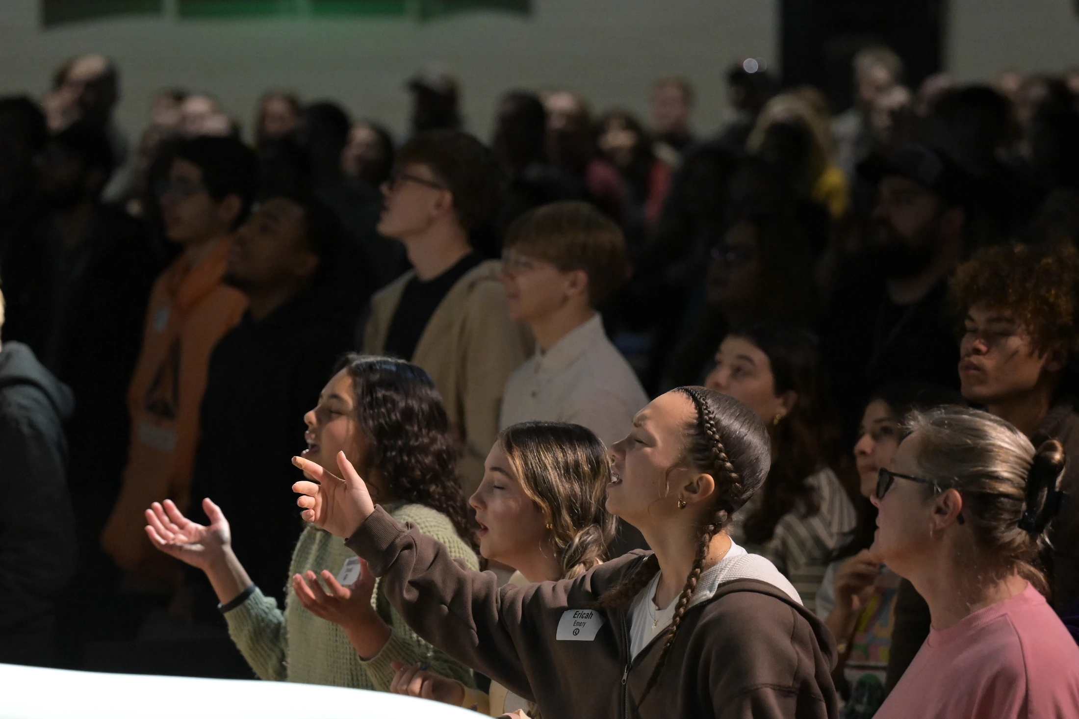 A diverse crowd of people stands together in a dimly lit room. In the foreground, several young women are singing with their hands raised in worship.
