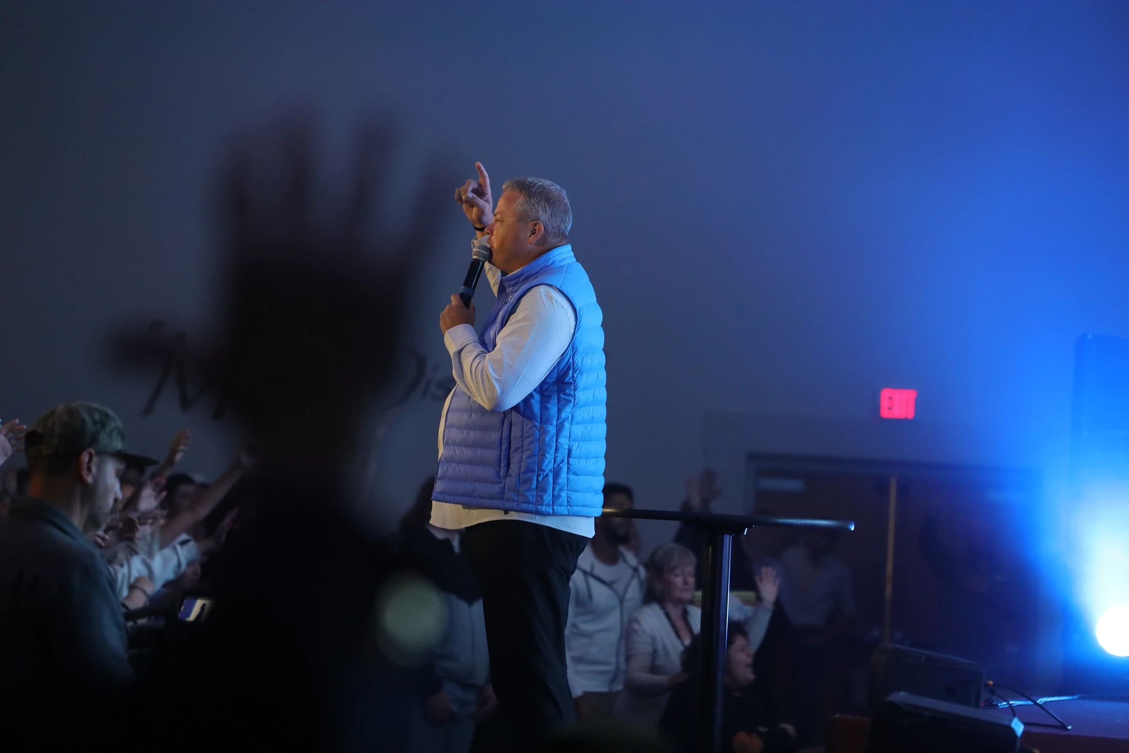 A man in a blue vest speaks into a microphone on a blue-lit stage, gesturing upwards. In front of him, a crowd watches, with some people raising their hands.