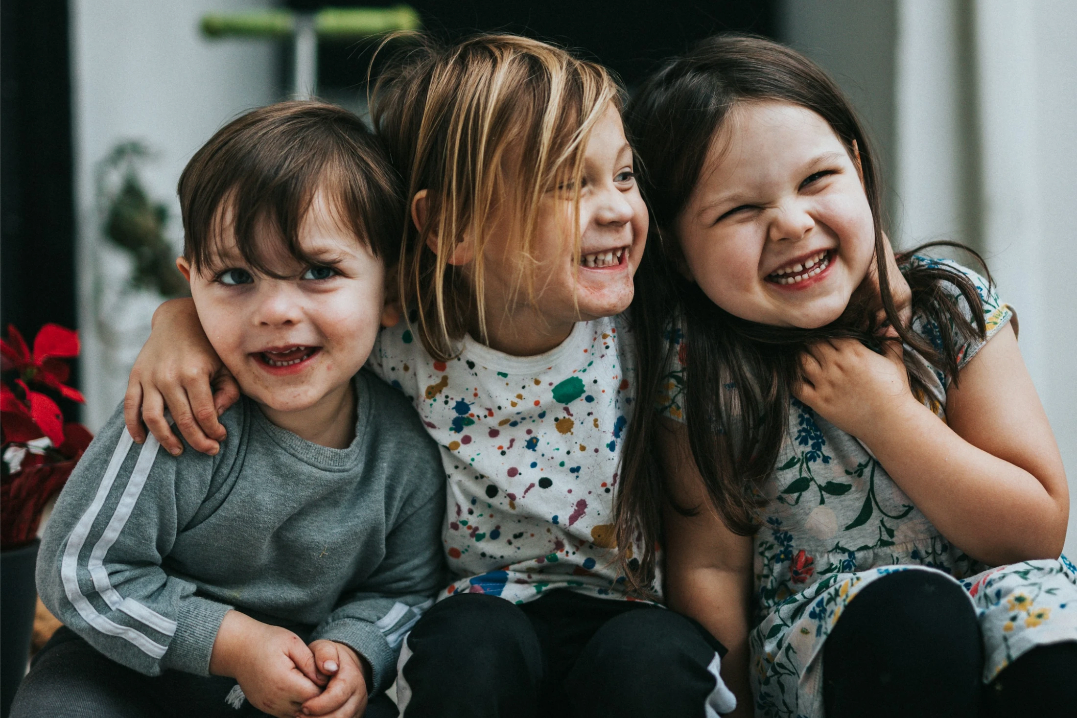 Three young children sit closely together, hugging and laughing joyfully. The middle child has their arm around the others in a happy, candid moment.