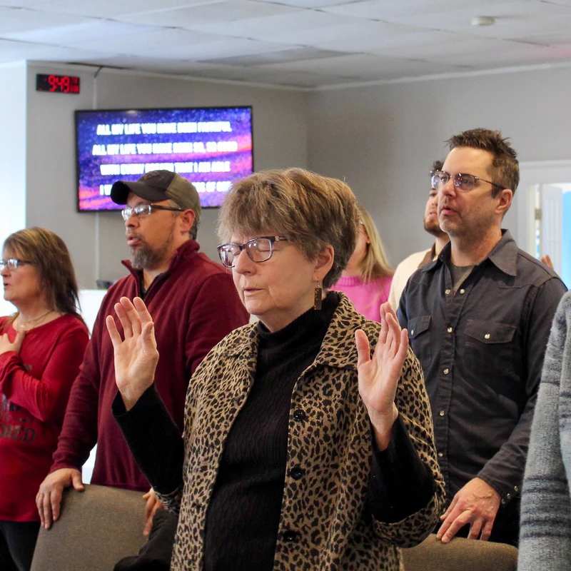 A diverse group of people stand together during a church service. Many are singing, and a woman in the center raises her hands in worship.