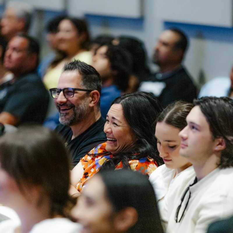 A diverse group of people sitting in an audience. In the center, a man with glasses smiles and a woman in a colorful blouse laughs while watching.