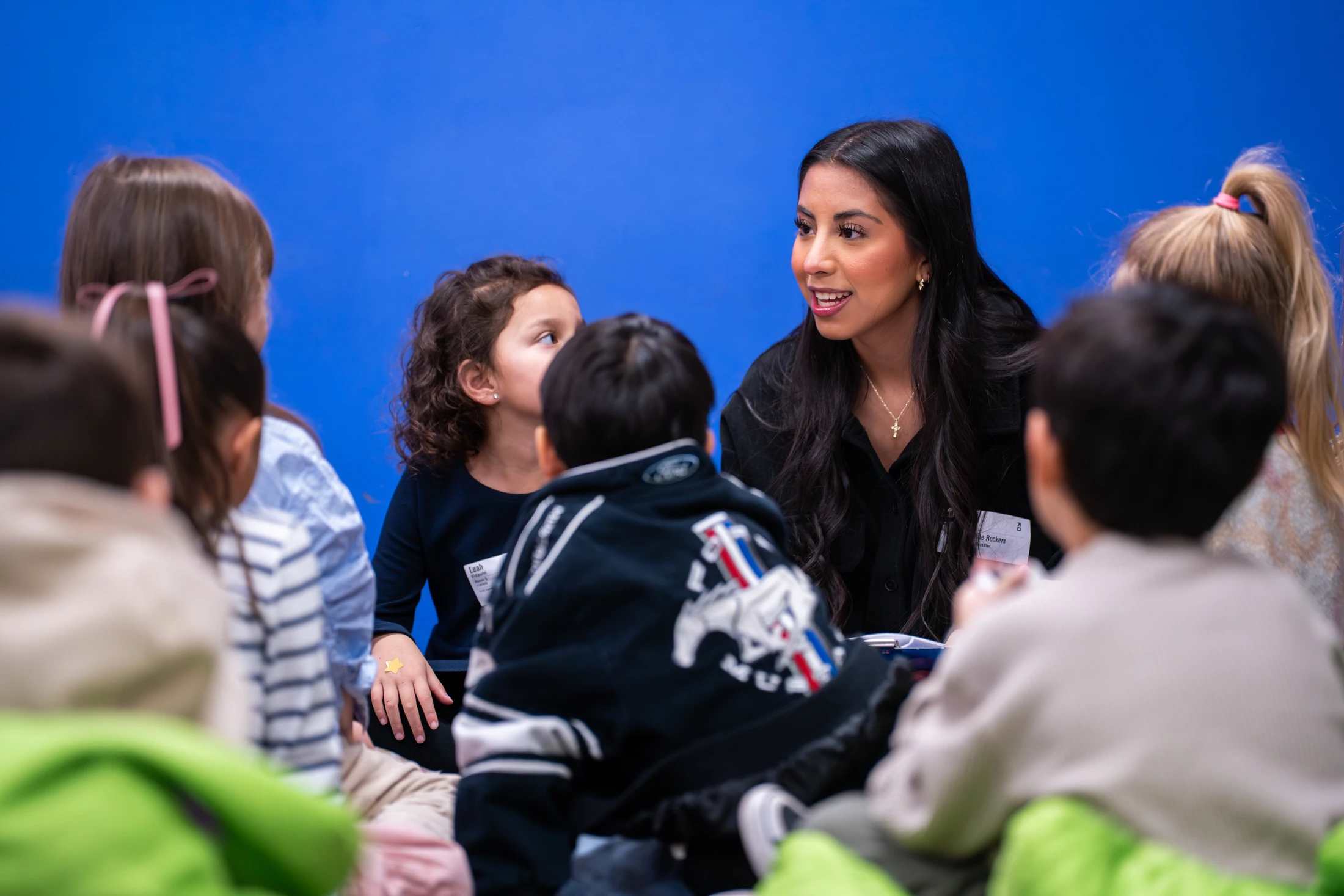 A woman with long dark hair sits in a circle on the floor with several young children, talking with them against a solid bright blue background.