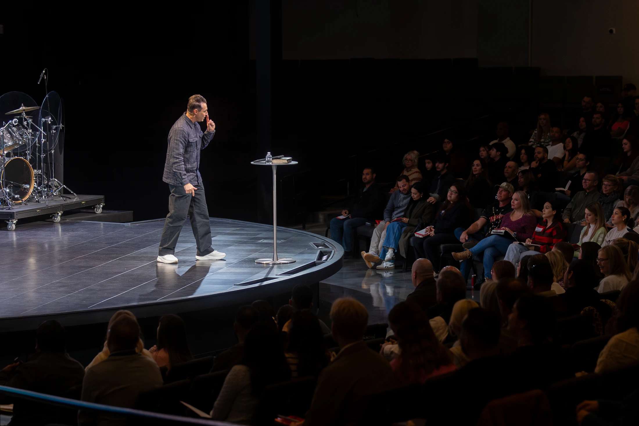 A man in a grey shirt stands on a dark, circular stage, speaking to an audience. He holds one finger to his lips in a shushing gesture.