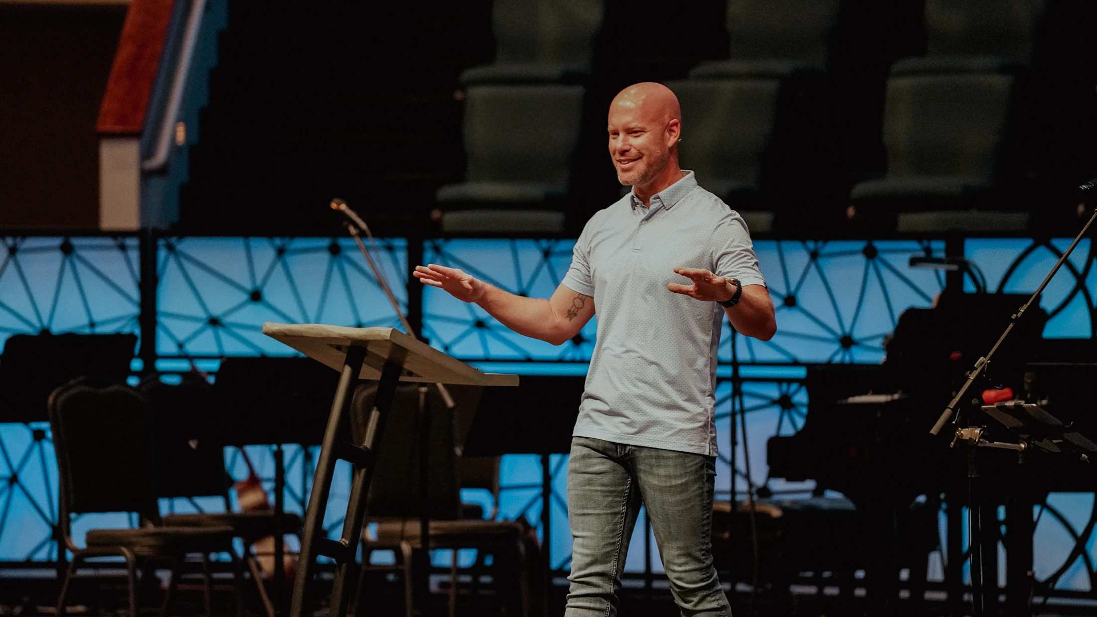 A smiling bald man in a light blue polo shirt and jeans stands on a stage, gesturing with his hands as he speaks from behind a lectern.